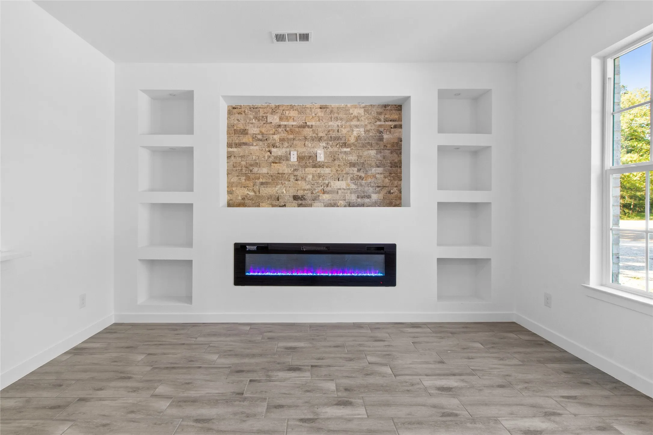 Modern entertainment space featuring custom built-in shelving flanking a brick accent wall, linear electric fireplace with blue LED accent lighting, wood-finish plank flooring, and abundant natural light from large picture window