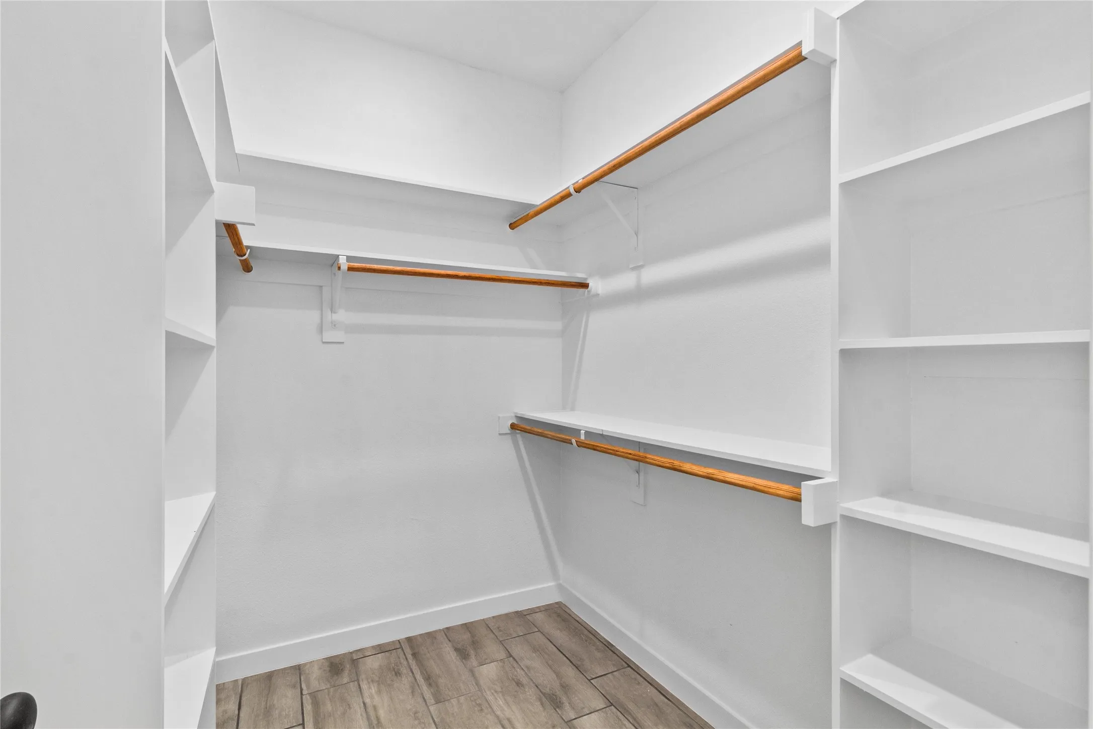 Spacious walk-in closet featuring custom wood-finish hanging rods, white built-in shelving throughout, and wood-finish flooring