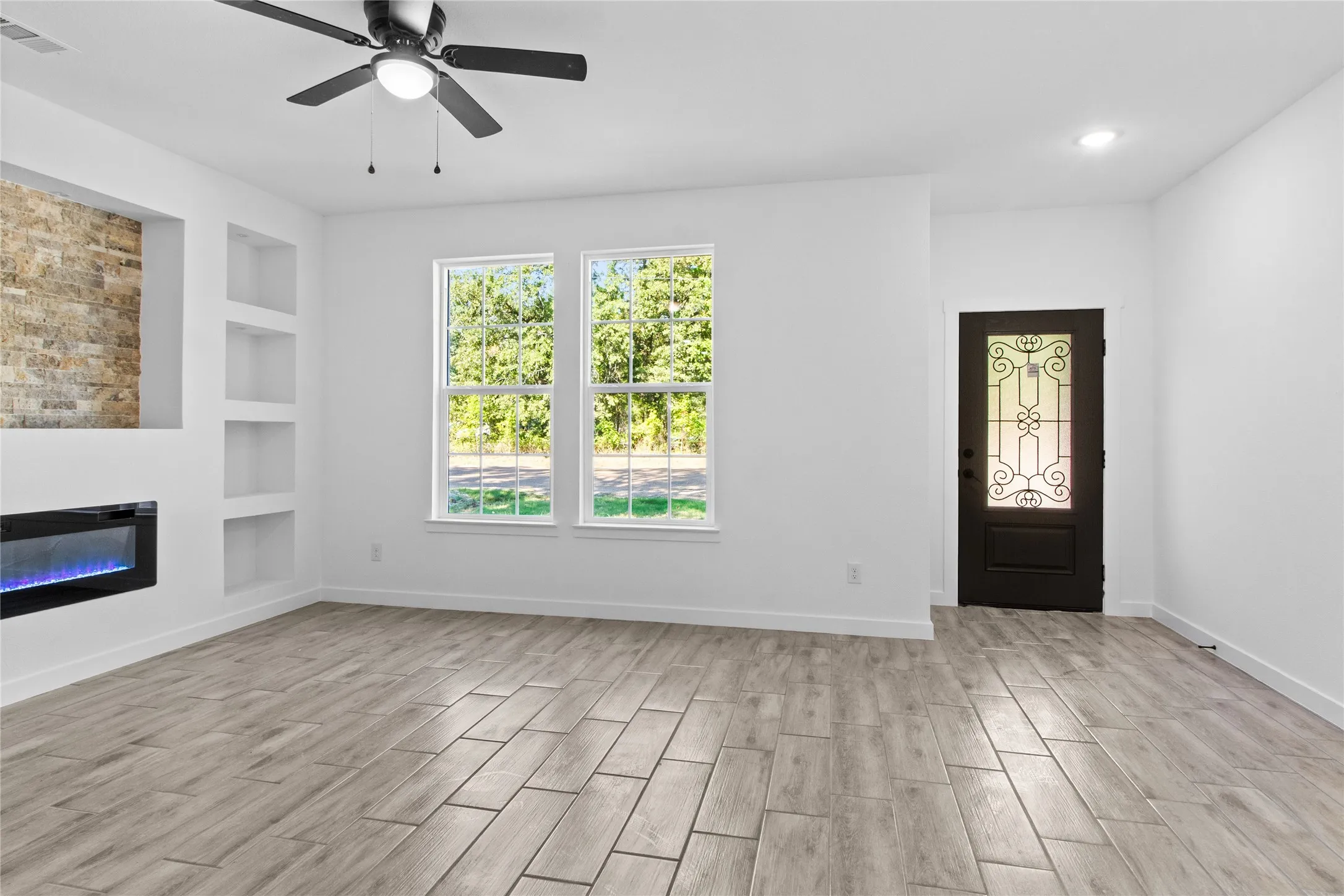 Wood-finish tile flooring throughout, featuring a built-in fireplace with stone-finish accent, multiple recessed shelving niches, a ceiling fan, and a decorative front entry door