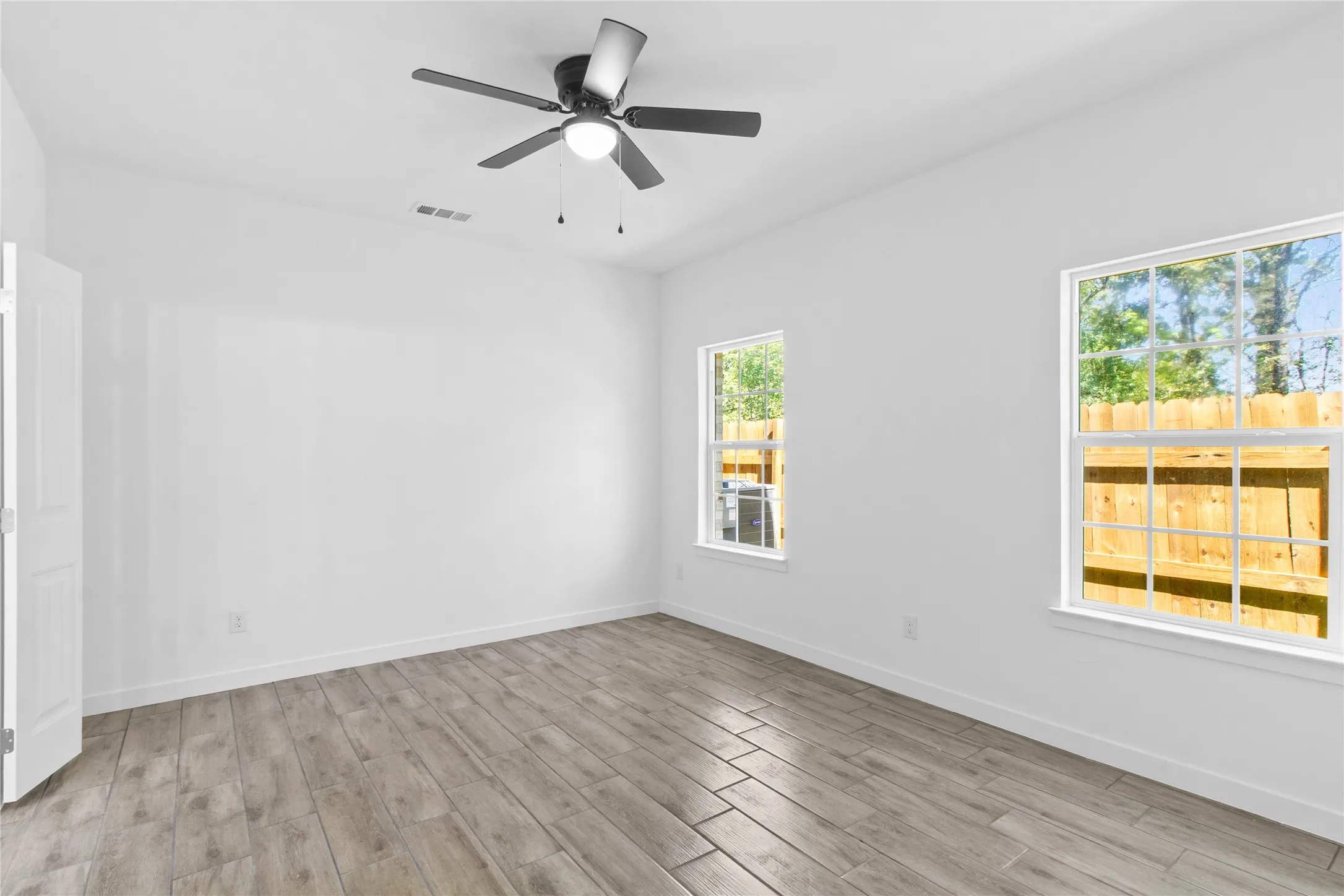 White walls, wood-finish flooring, two window openings, and a ceiling fan with integrated lighting