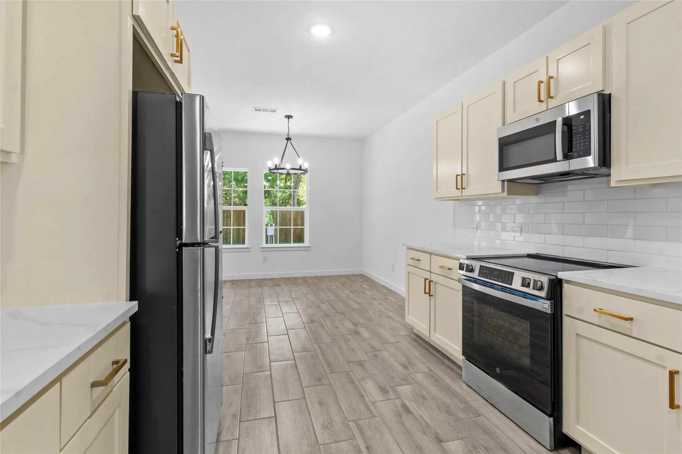Galley-style kitchen featuring cream-colored cabinetry with brass hardware, stainless steel appliances including a French-door refrigerator and built-in microwave, subway tile backsplash, wood-finish flooring, and abundant natural light from dual windows