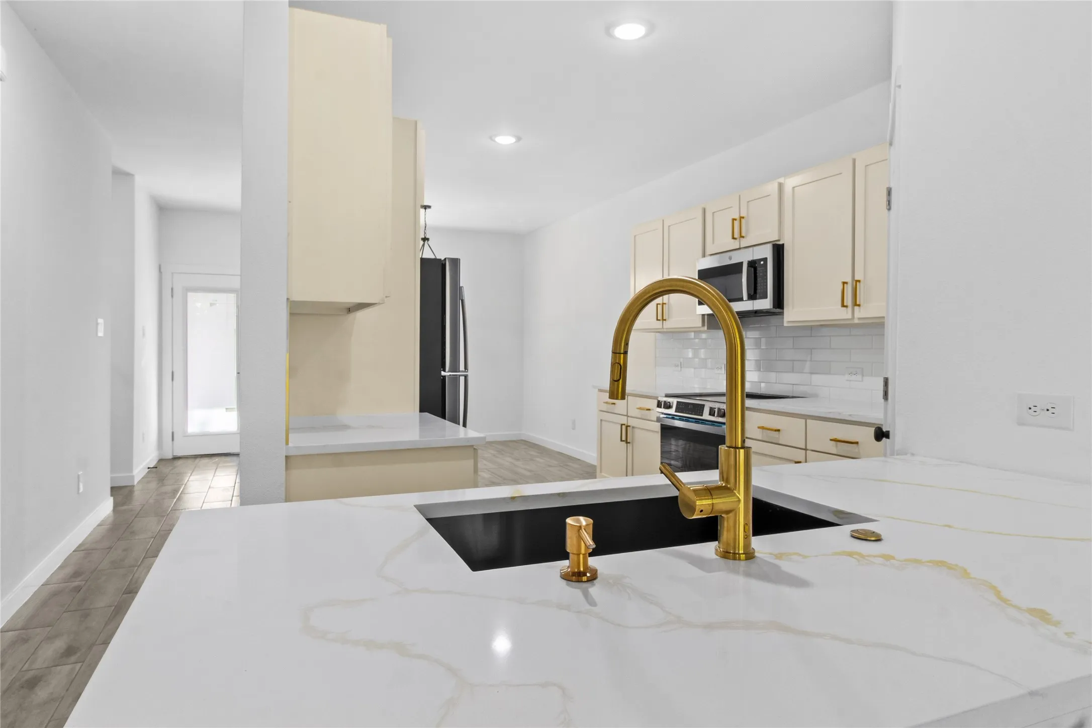 Expansive kitchen island with a dark undermount sink and gold-tone faucet, complemented by light-toned cabinetry and a white subway tile backsplash, and recessed lighting