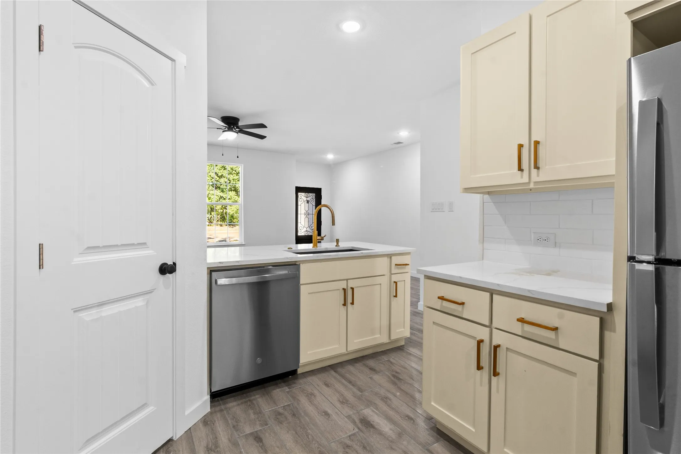Modern kitchen featuring cream-colored cabinetry with brushed brass hardware, stainless steel appliances including a dishwasher and refrigerator, white subway tile backsplash, wood-finish flooring, recessed ceiling lighting, and a ceiling fan