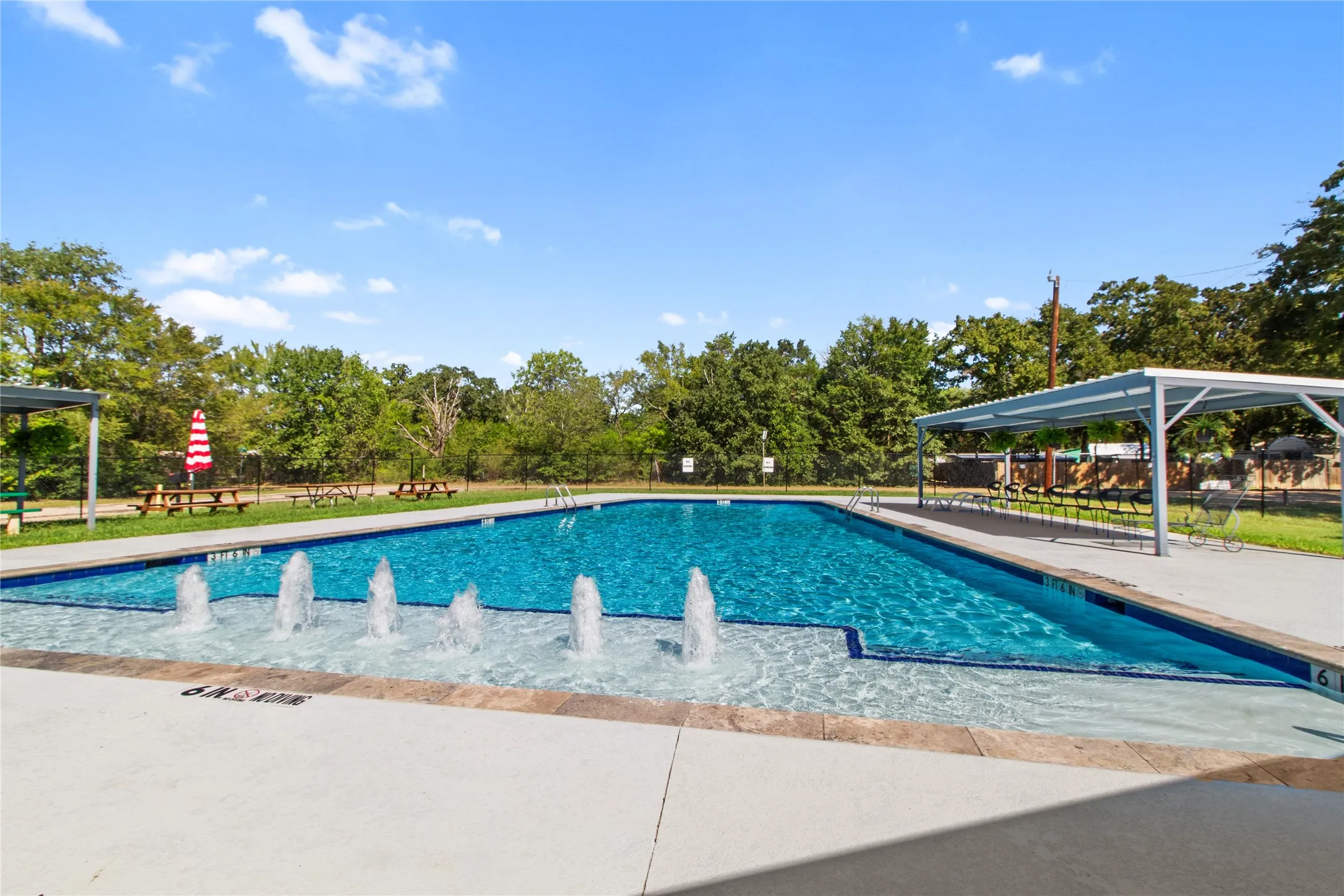 Resort-style swimming pool featuring crystal-clear water, multiple water fountains, concrete decking, and overhead shade structures