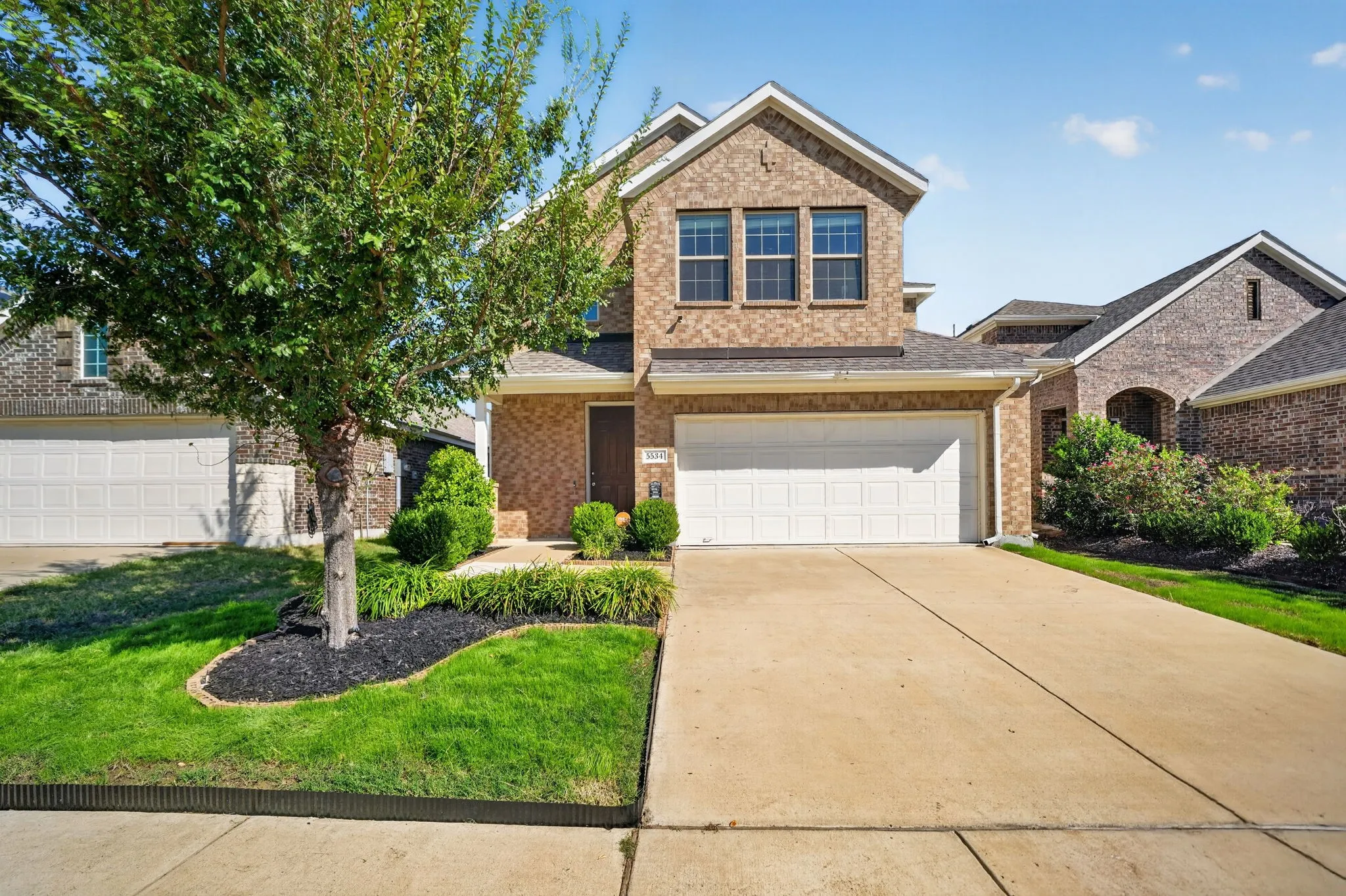 Brick exterior with an attached two-car garage, concrete driveway, and front lawn with mature tree and landscaping