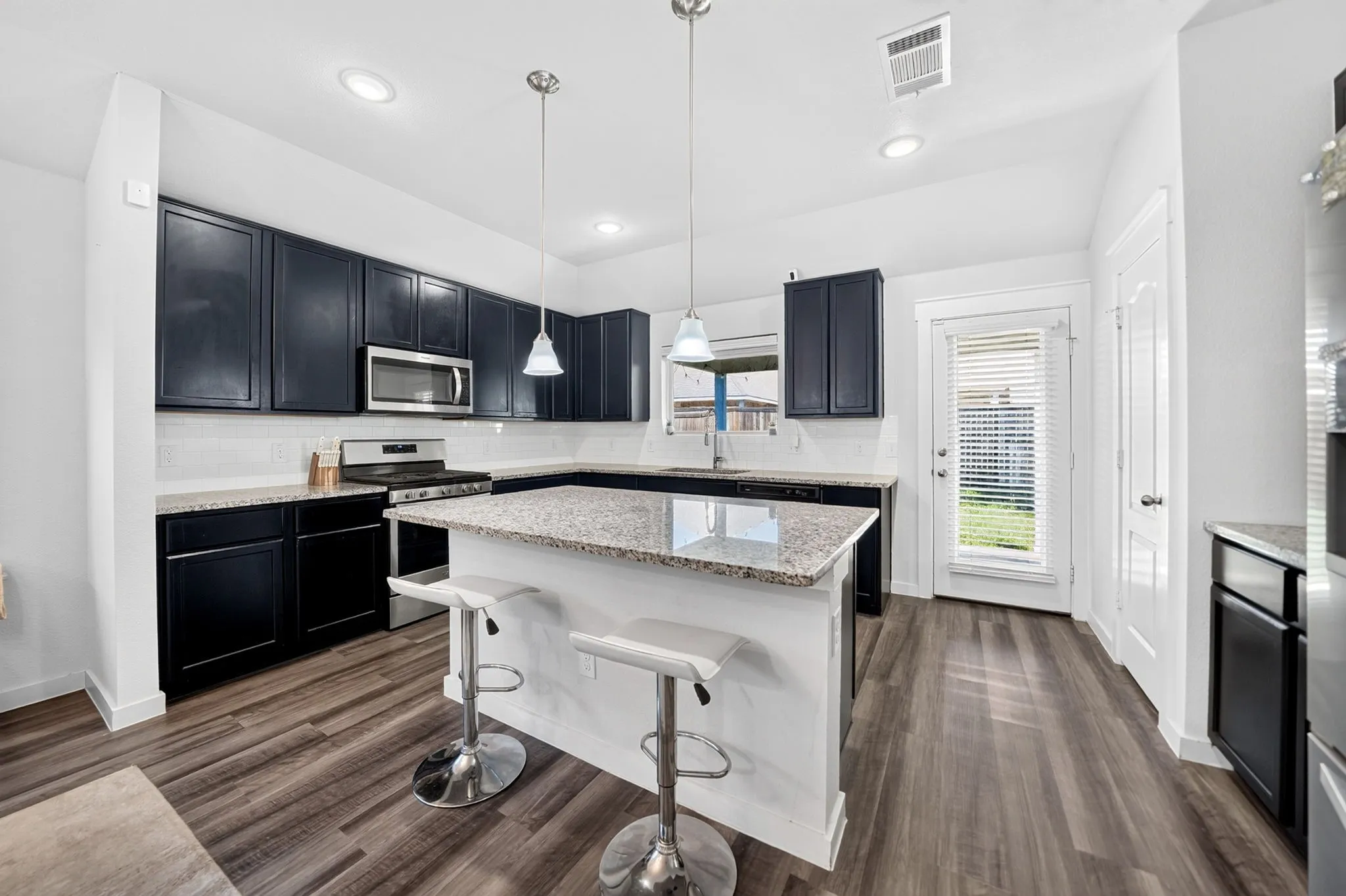 Modern kitchen featuring dark cabinetry with stainless steel appliances, white quartz countertops, a center island with seating, pendant lighting, and wood-finish flooring throughout the open layout