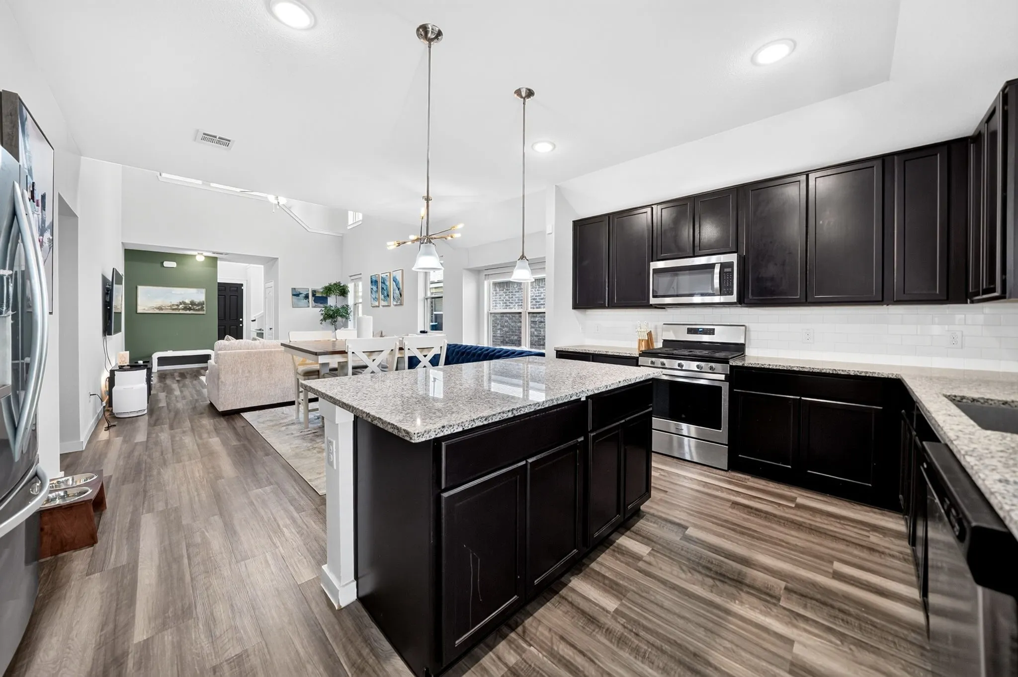 Open-concept kitchen featuring dark wood-finish cabinetry, light-toned granite countertops, a subway tile backsplash, stainless steel appliances, and wood-finish flooring