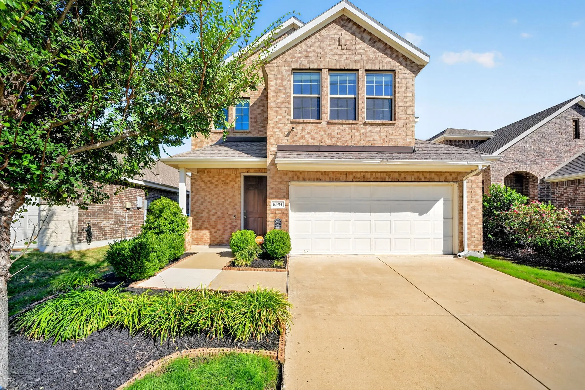 Charming brick two-story home featuring a clean concrete driveway, white garage door, and manicured front landscaping with ornamental grasses and mature greenery
