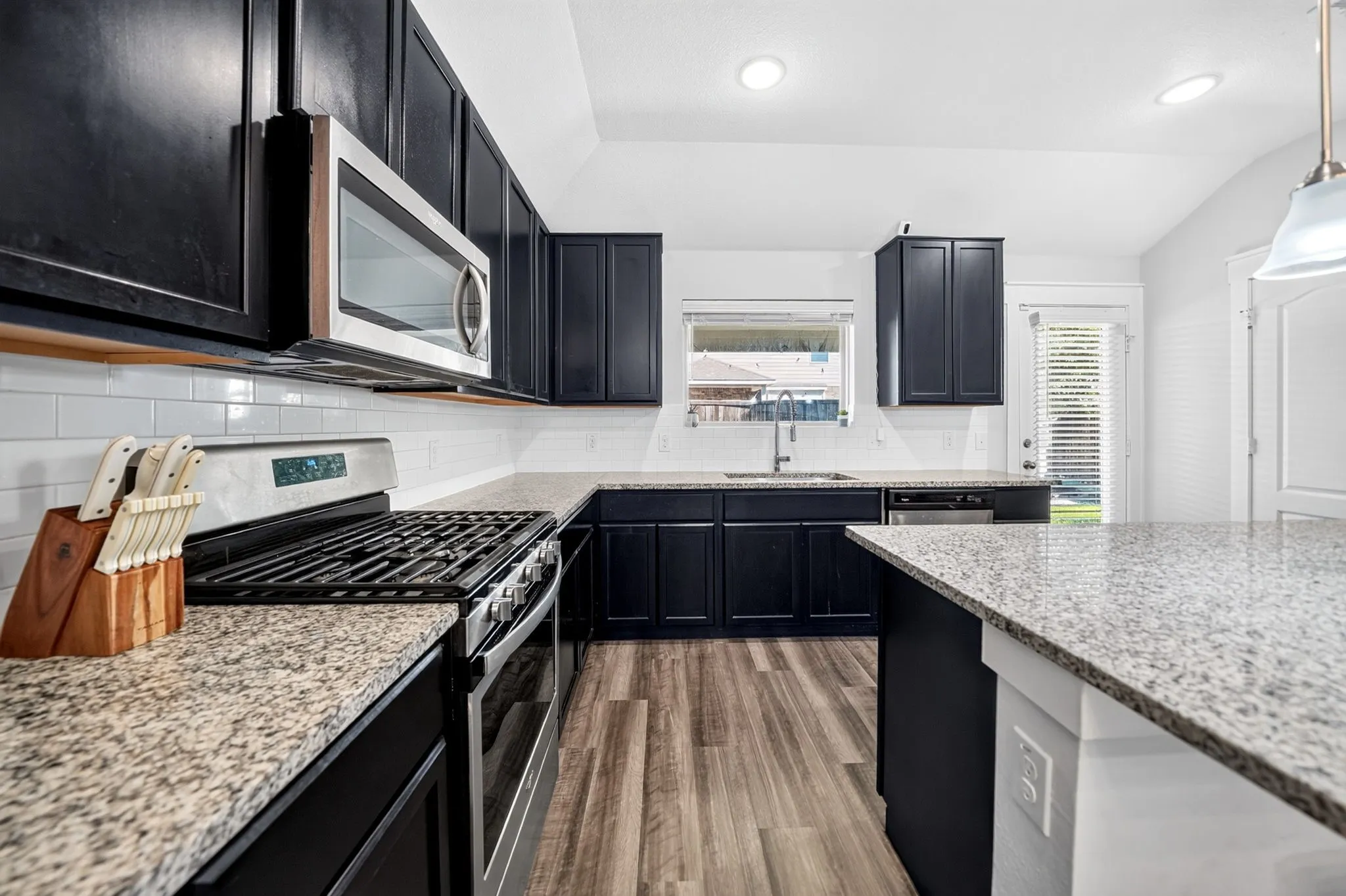 Modern kitchen featuring sleek black cabinetry with stainless steel gas range, granite-style countertops, subway tile backsplash, recessed ceiling lighting, and wood-finish flooring throughout the open layout