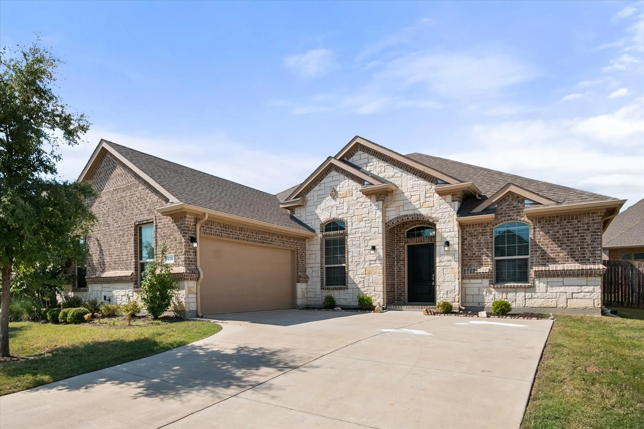 Brick and stone facade, arched entryway, attached garage, concrete driveway, and established landscaping