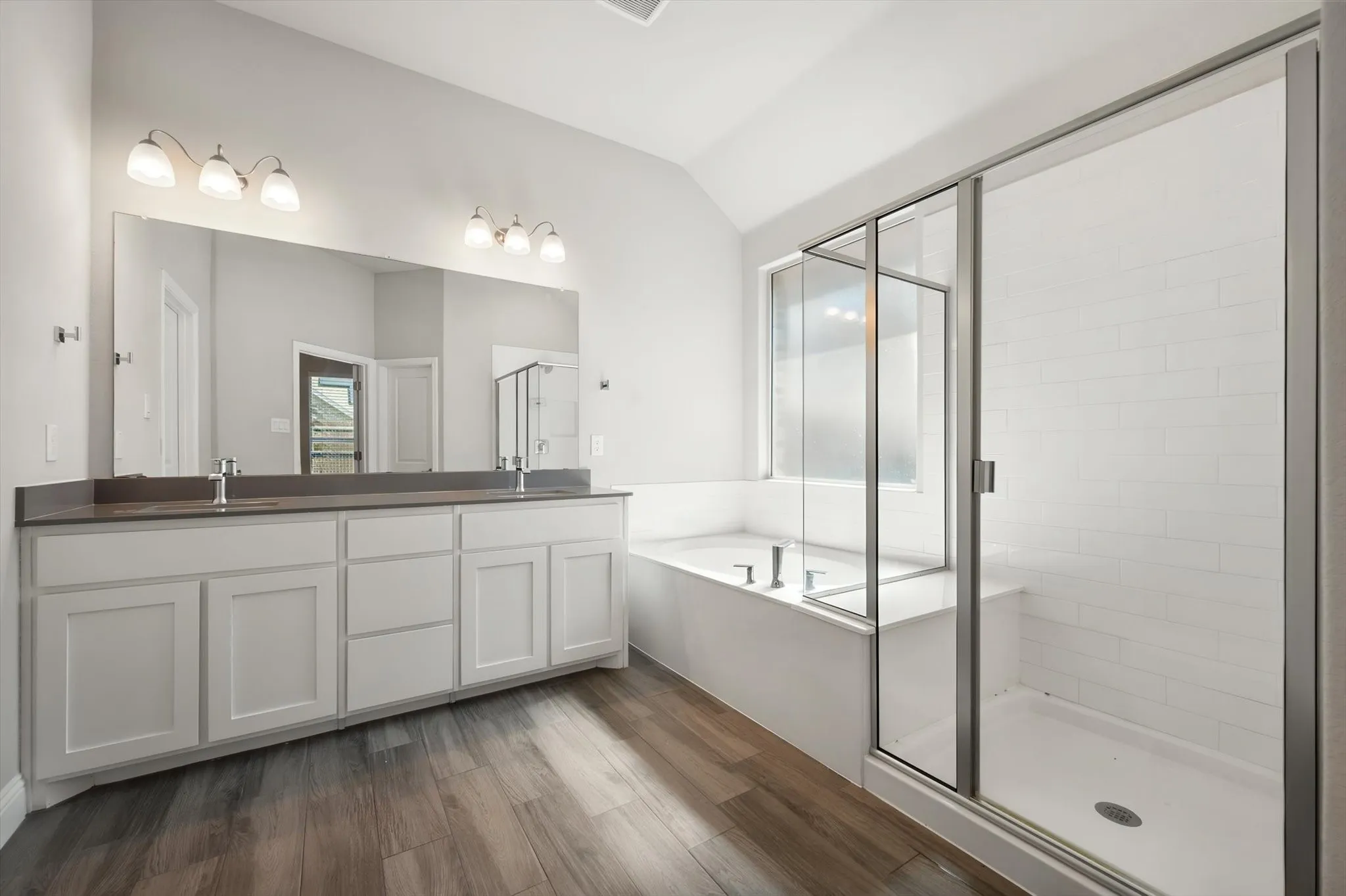 Wood-finish flooring, dual vanity with white cabinetry, built-in bathtub, glass-enclosed shower, and subway tile shower surround
