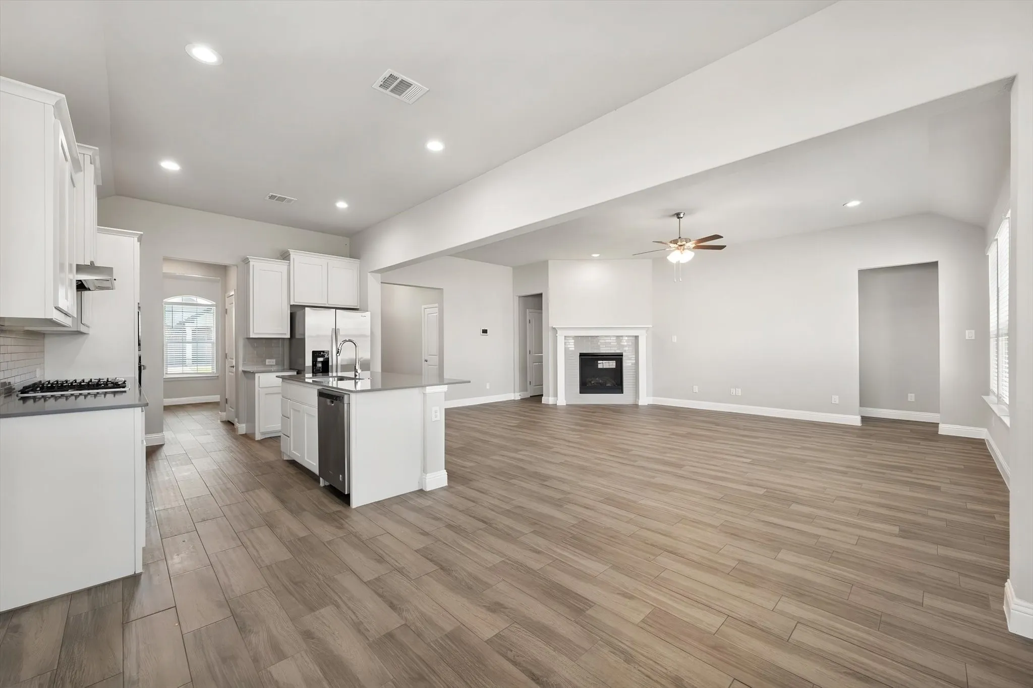 Open-concept living area featuring wood-finish flooring, a kitchen island with sink, white shaker cabinetry, stainless steel appliances, and a built-in fireplace with mantel