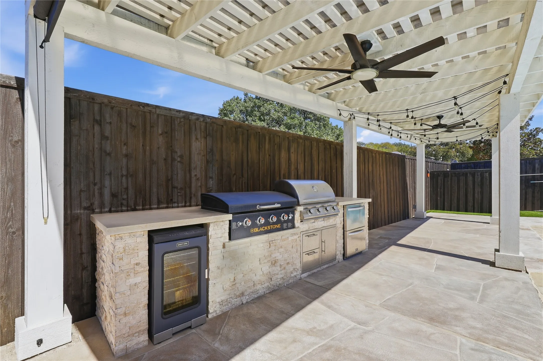 Expansive outdoor patio featuring a slatted pergola, integrated outdoor kitchen with built-in grill and refrigerator, multiple ceiling fans, and string lighting