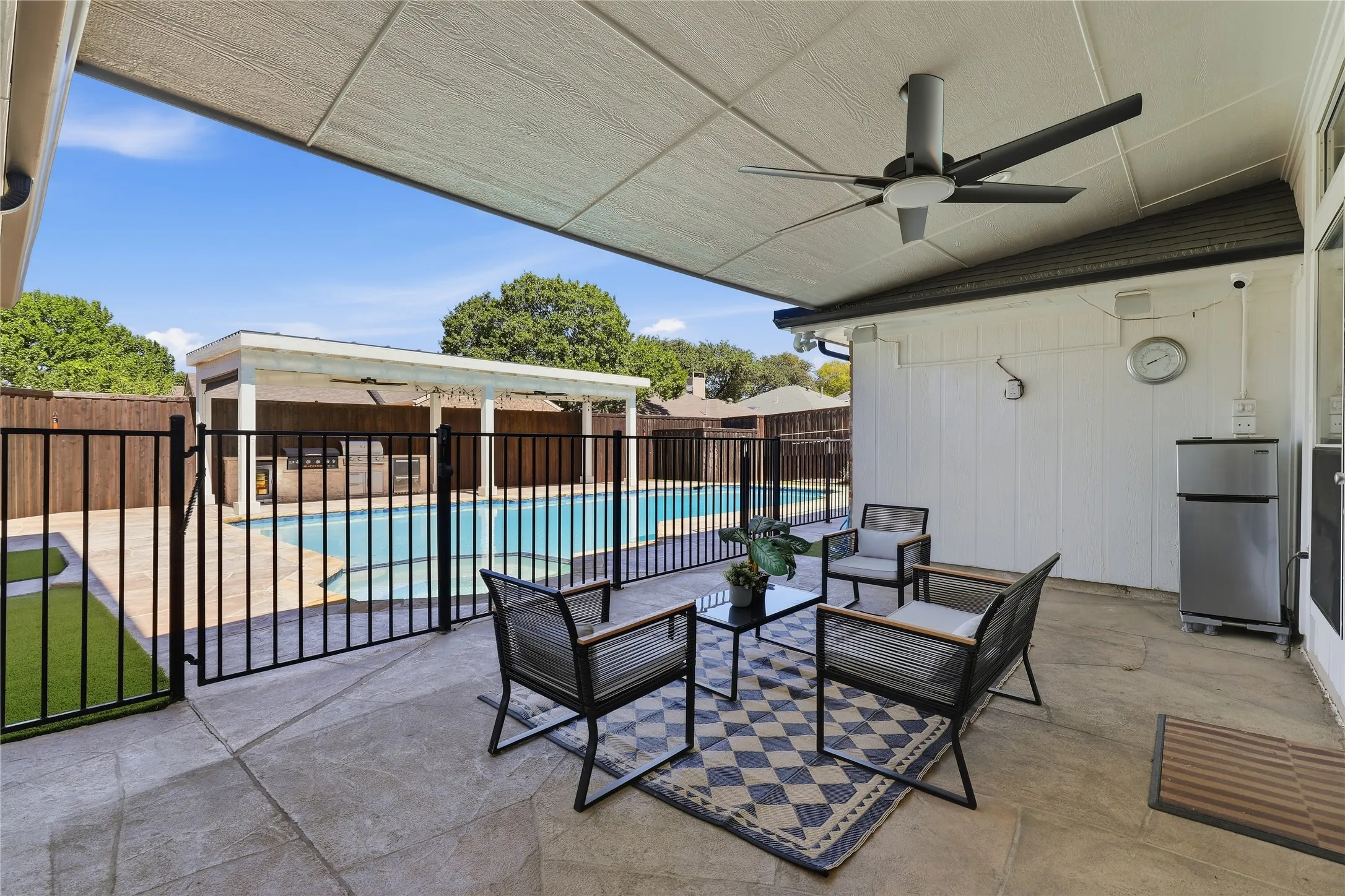 Covered patio with a ceiling fan, concrete flooring, and a black metal fence enclosing a swimming pool with a paved surround and an adjacent outdoor structure