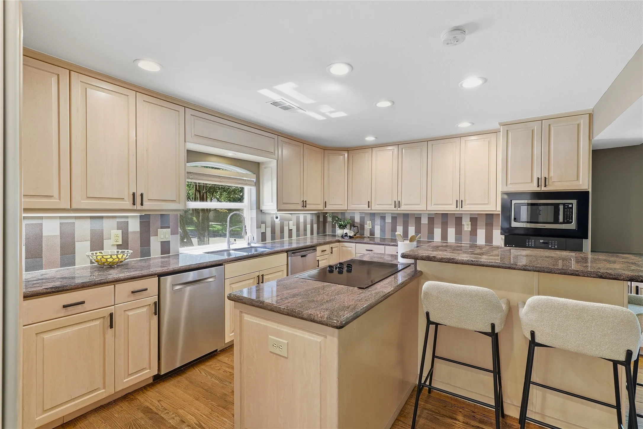 Kitchen featuring wood-finish flooring, light wood cabinetry, stone countertops, a tile backsplash, and stainless steel appliances