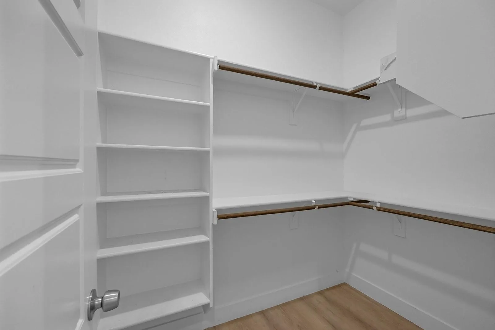 Spacious walk-in closet featuring built-in shelving along one wall, dual wooden hanging rods at varying heights, and wood-finish flooring for organized storage and accessibility