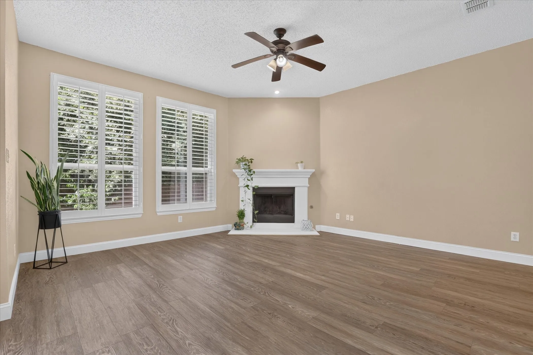 Wood-finish flooring throughout, featuring a white mantle fireplace, multiple windows with white louvered blinds, and a ceiling fan with integrated lighting