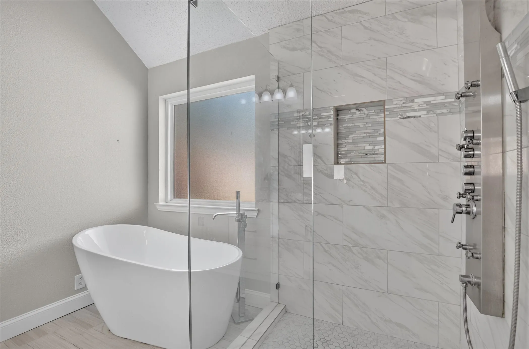 Freestanding bathtub with floor-mounted faucet, glass-enclosed shower with tile surround, mosaic tile accent, recessed shower niche, and multi-function shower panel