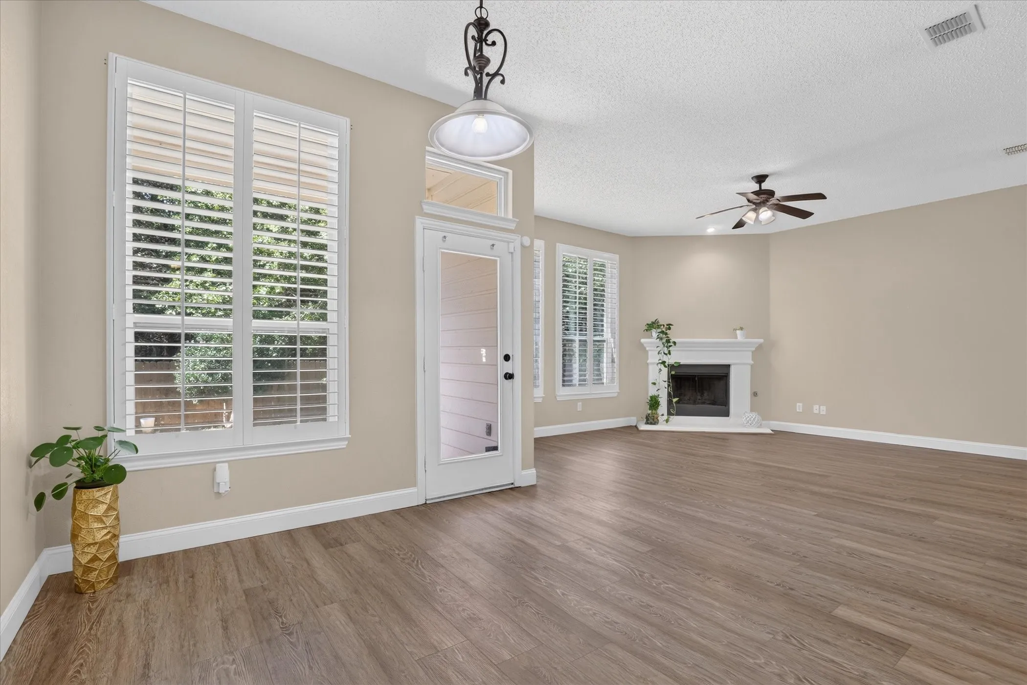 Wood-finish flooring, white window shutters, white-framed glass paneled door, ceiling fan, and white fireplace surround