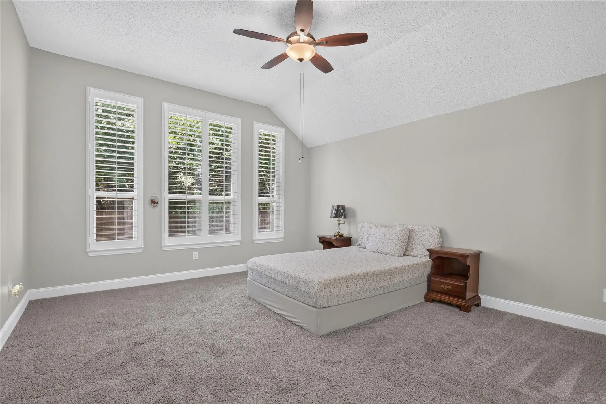 Vaulted ceiling with a ceiling fan, three tall windows with louvered blinds, and gray carpeting