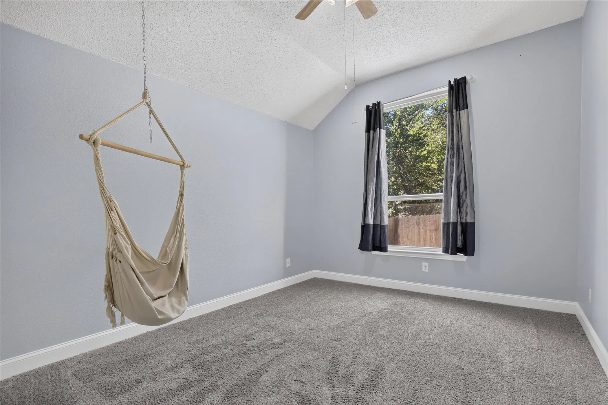 Vaulted ceiling, ceiling fan, window with exterior landscaping, light gray walls, and gray carpeted flooring