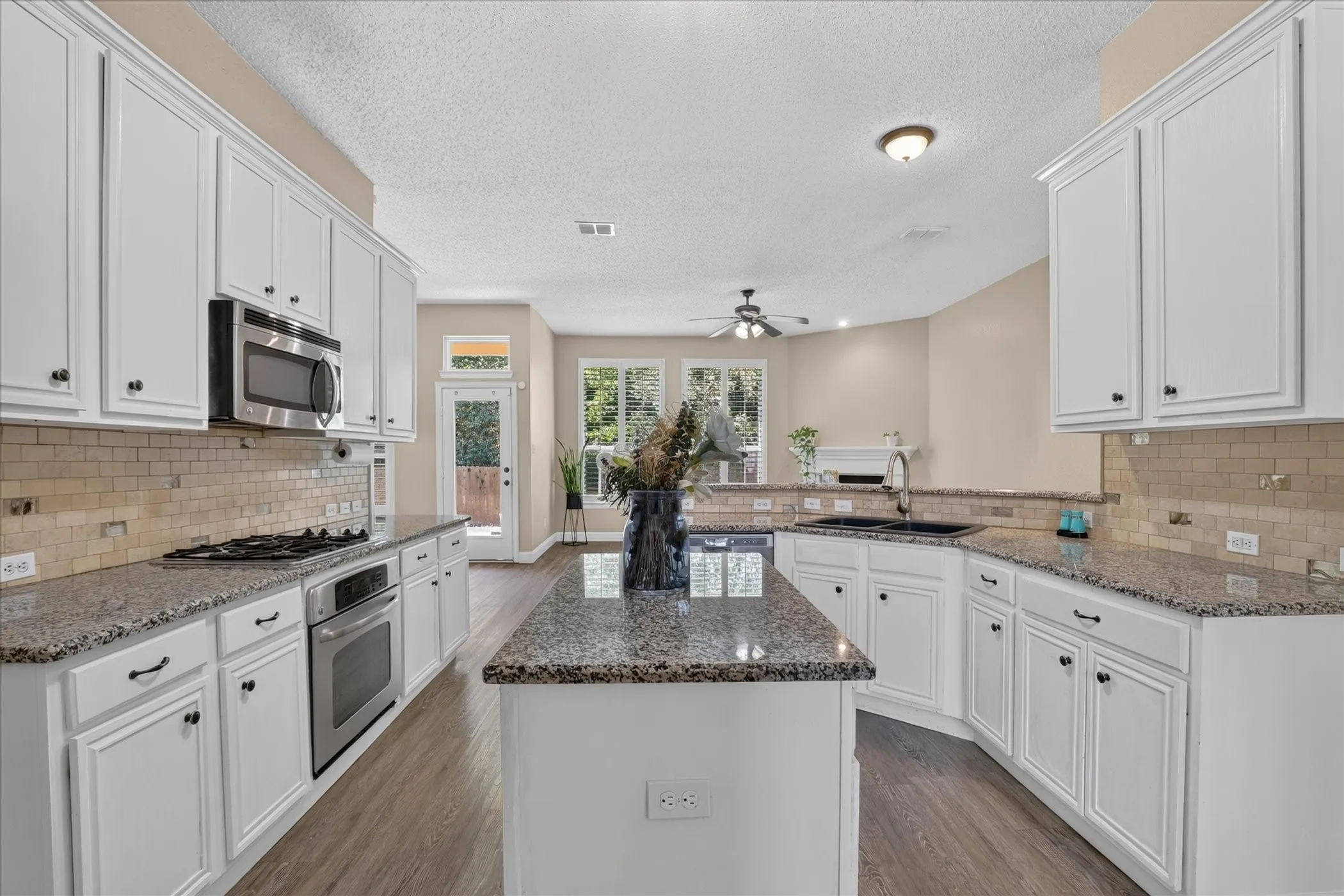 White cabinetry, speckled countertops, stainless steel appliances, tile backsplash, and wood-finish flooring
