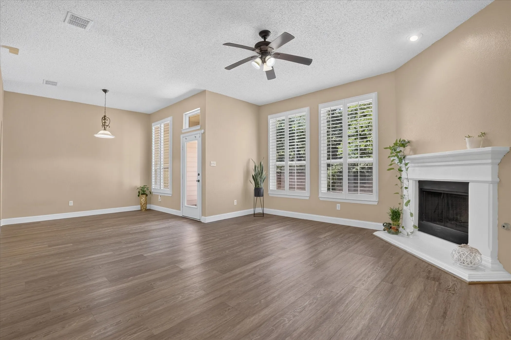 Spacious room featuring wood-finish flooring, a ceiling fan, a white fireplace with a hearth, and multiple windows with white plantation shutters