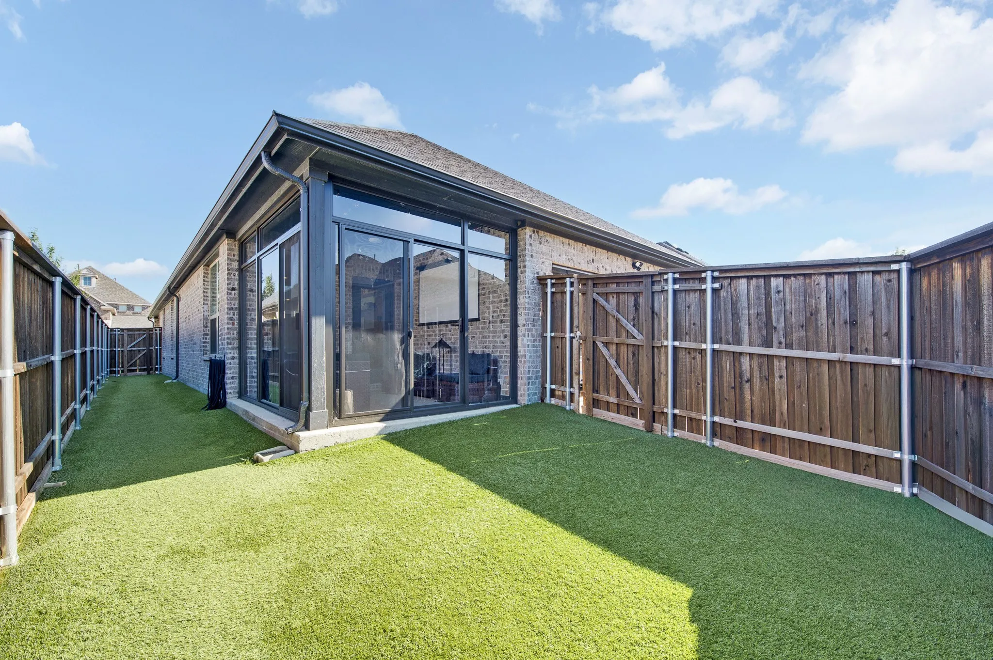 Modern backyard retreat featuring a spacious manicured lawn, contemporary glass-paneled studio or garden room with dark framing and pitched roofline, horizontal wood-finish fencing with metal railings, and mature landscaping throughout