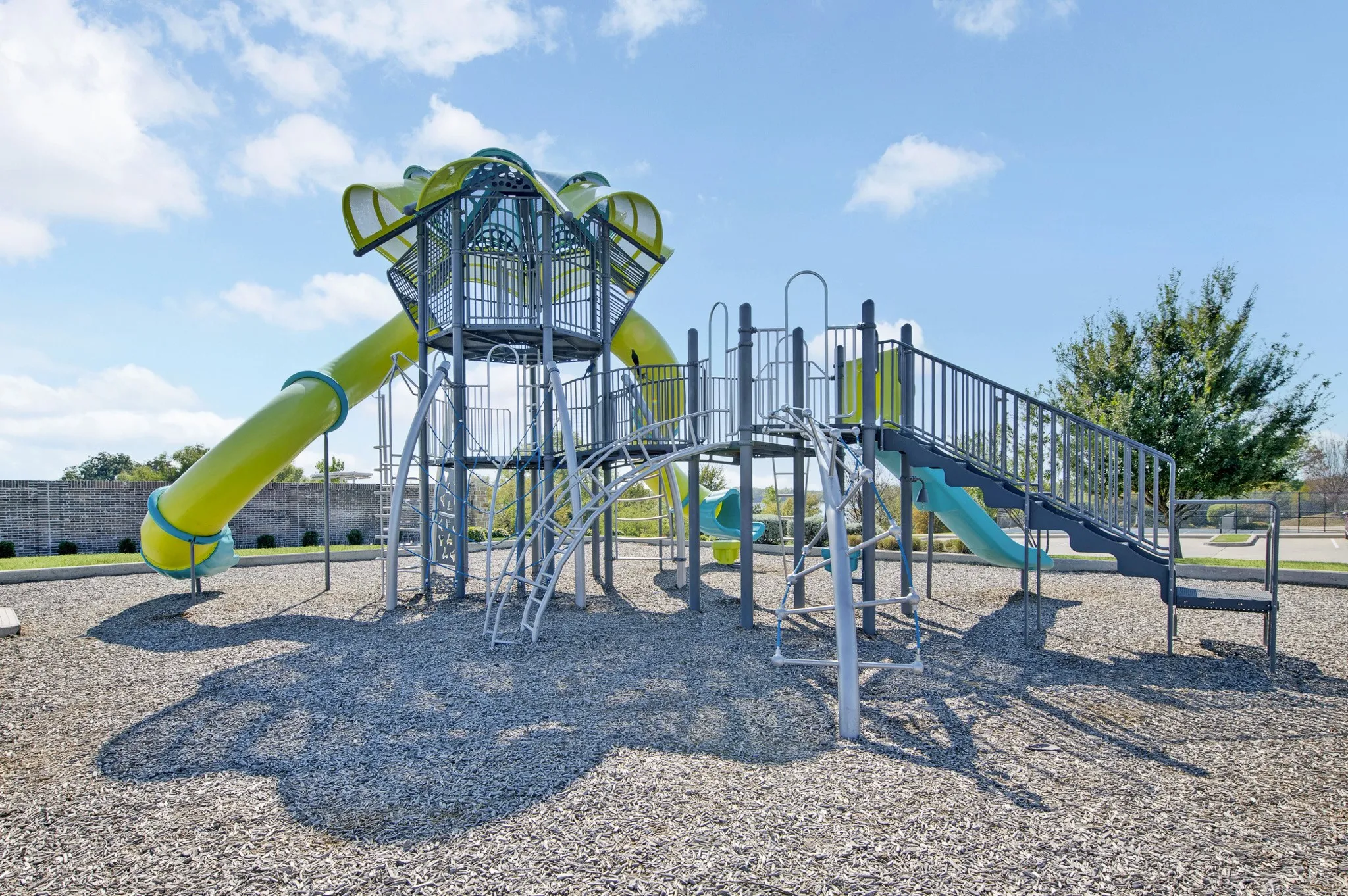 Modern playground featuring a tall spiral slide tower with multiple entry points, climbing structures, and ground-level play equipment, all set on a cushioned safety surface with mature landscaping and open green space surrounding the recreational area