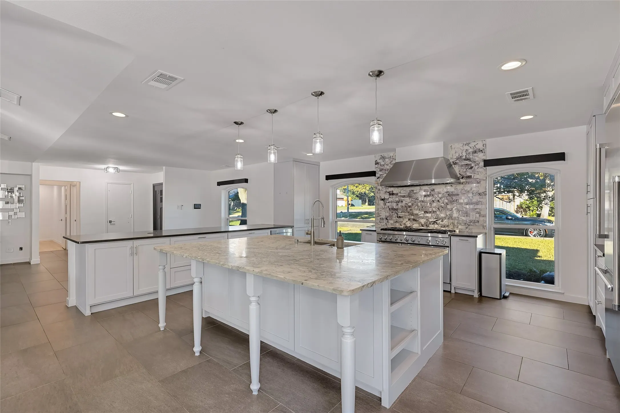 Spacious kitchen featuring an expansive central island with a light-toned countertop, white cabinetry with dark countertops, a stainless steel range hood, and tile flooring