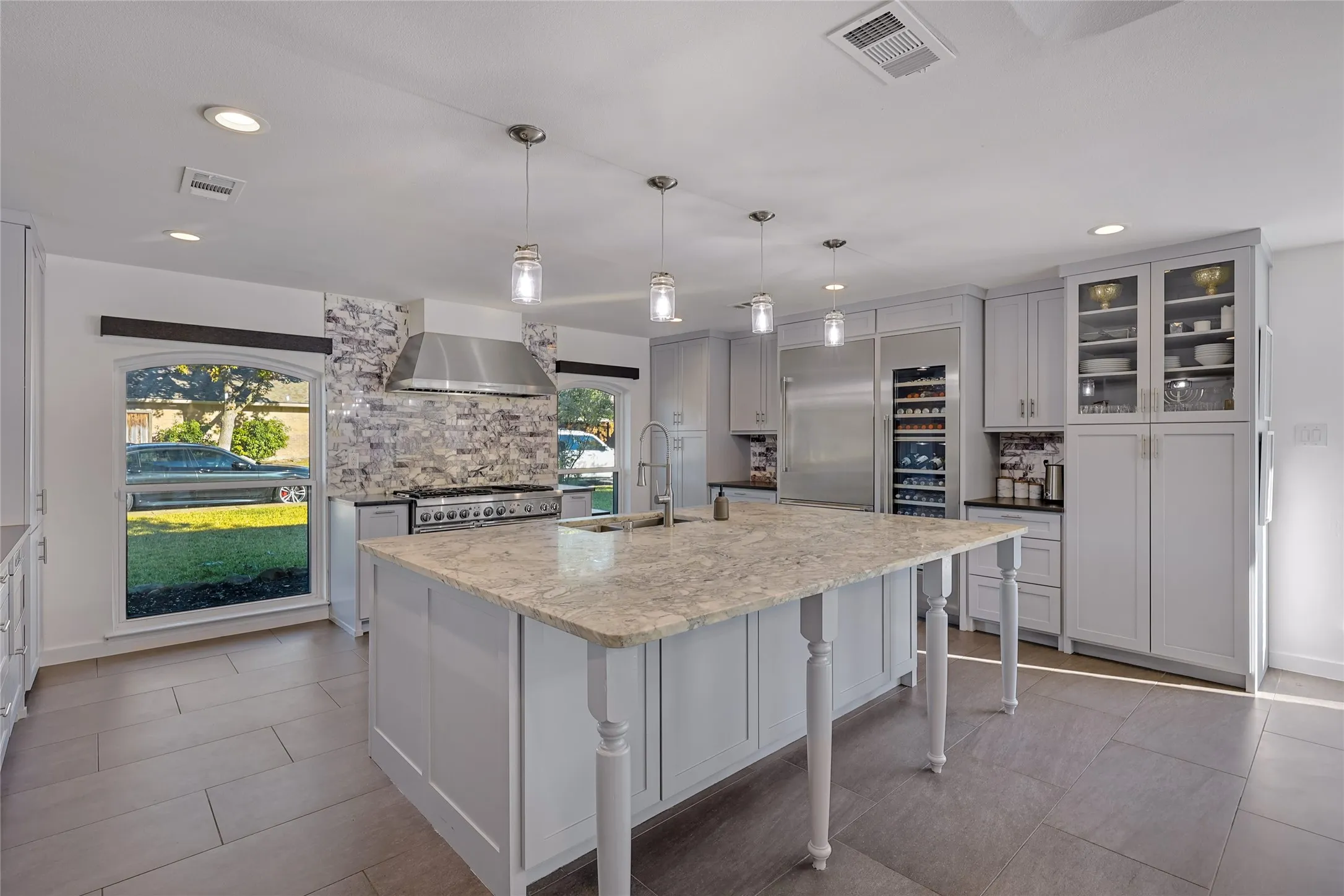 Kitchen featuring a central island with a light-toned countertop, stainless steel appliances, extensive cabinetry with some glass-front doors, recessed lighting, and a patterned tile backsplash