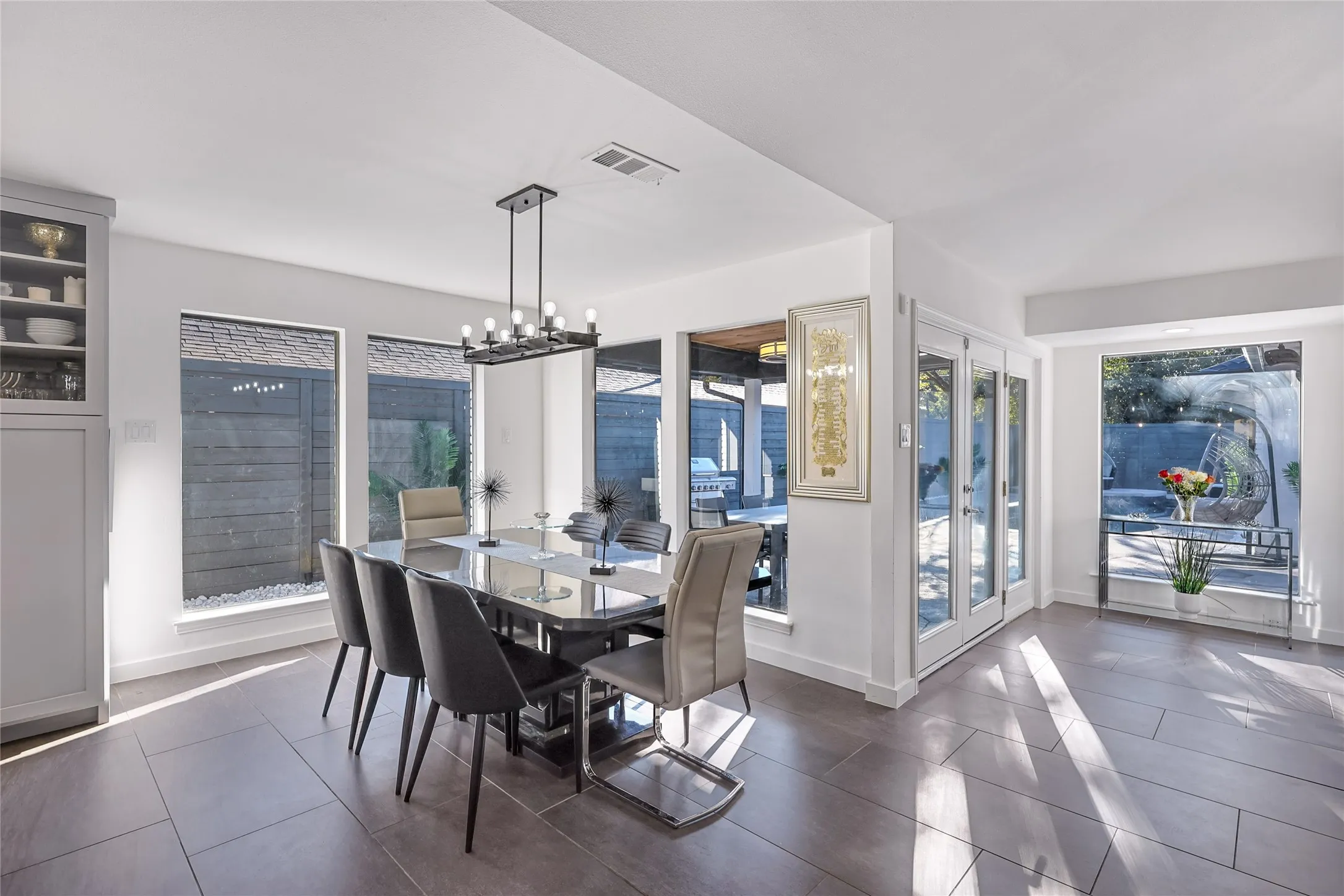 Modern dining area flooded with natural light through expansive glass doors and windows, featuring a dark wood-finish dining table, contemporary pendant lighting, and sleek dark-finish chairs