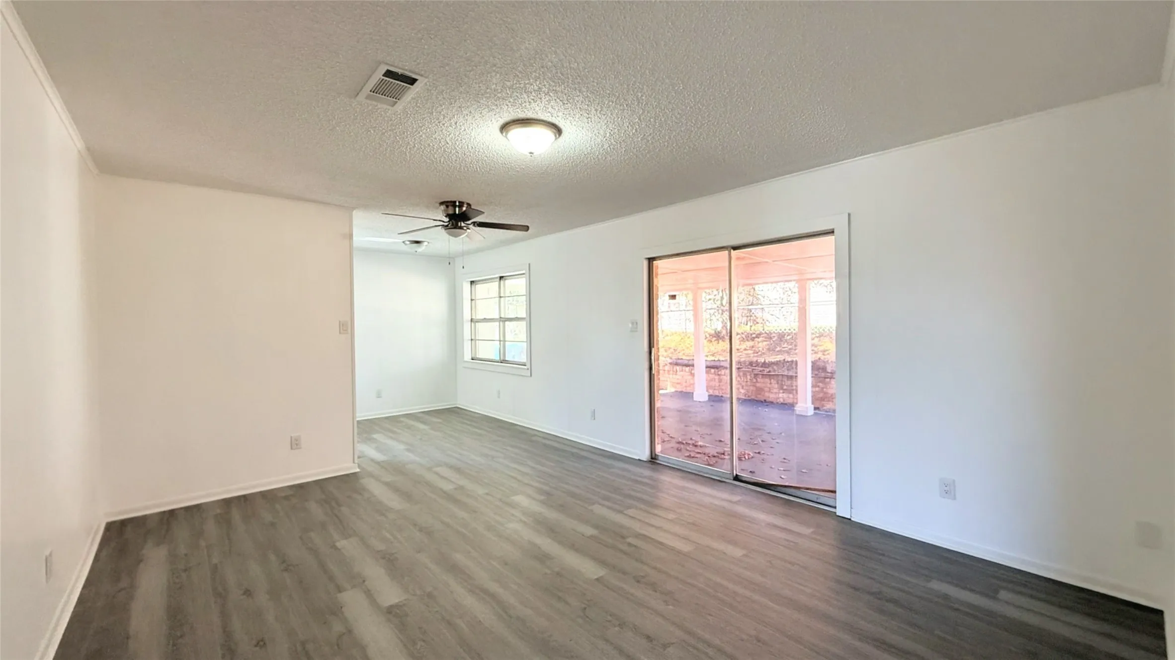 Wood-finish flooring throughout, sliding glass door leading to an outdoor area, ceiling-mounted light fixture, additional ceiling fan, and large window with grid pattern