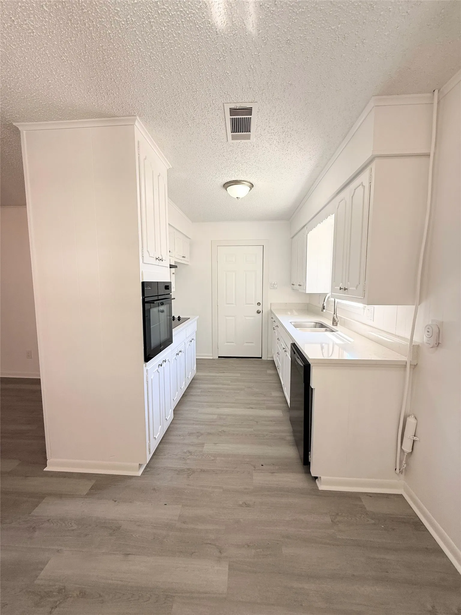 Galley kitchen featuring white cabinetry, light-toned wood-finish flooring, white countertops, built-in oven, and a black dishwasher
