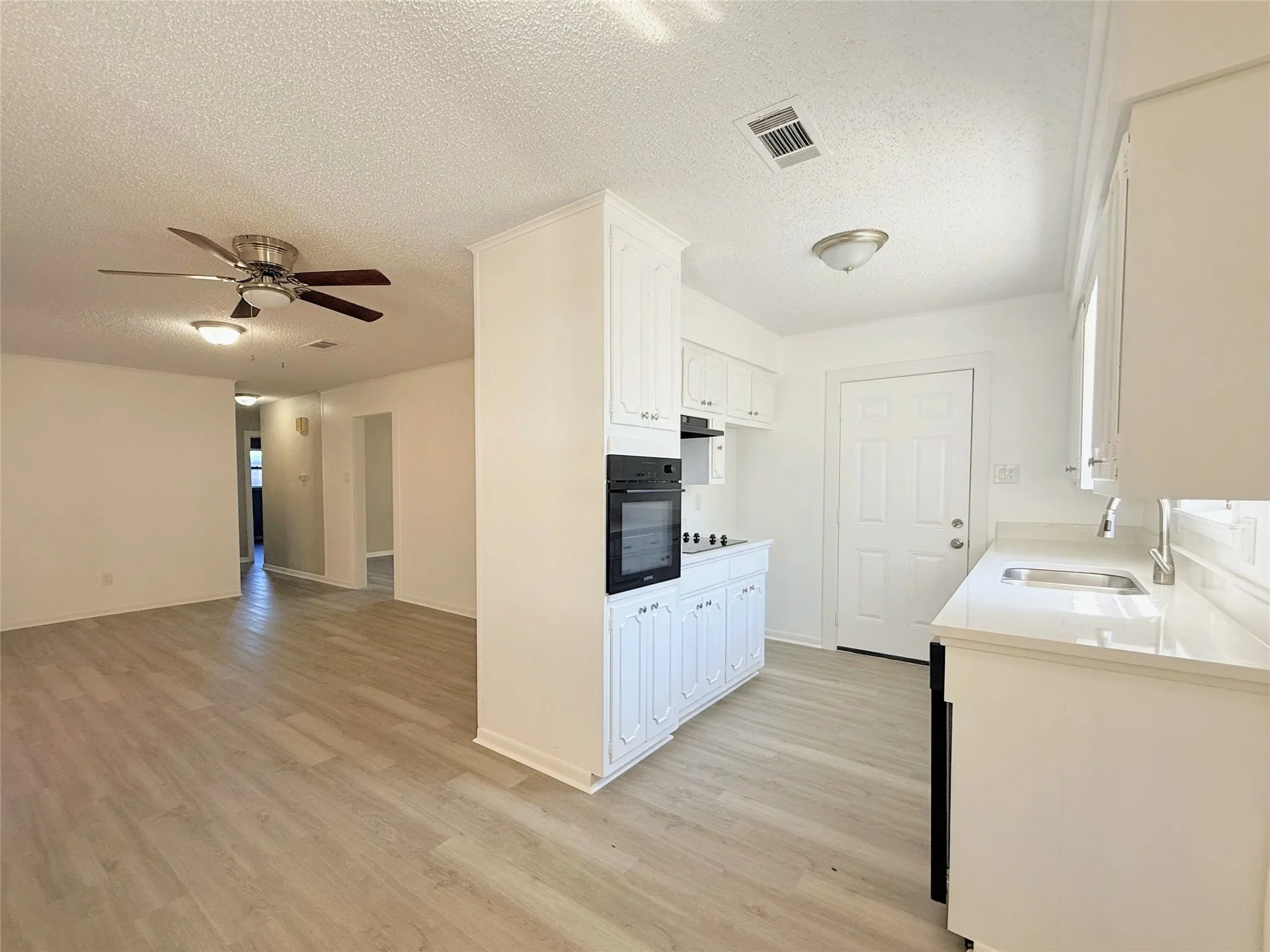Open-concept living area with wood-finish flooring, ceiling fan, white cabinetry, integrated oven, and a kitchen sink with a countertop