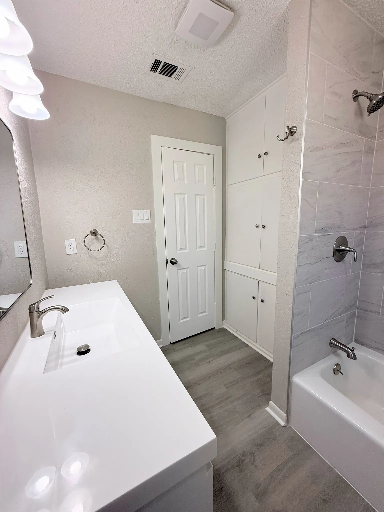 White bathtub with a brushed nickel showerhead and tiled surround, white vanity with an integrated sink and brushed nickel faucet, wood-finish flooring, and built-in white cabinetry