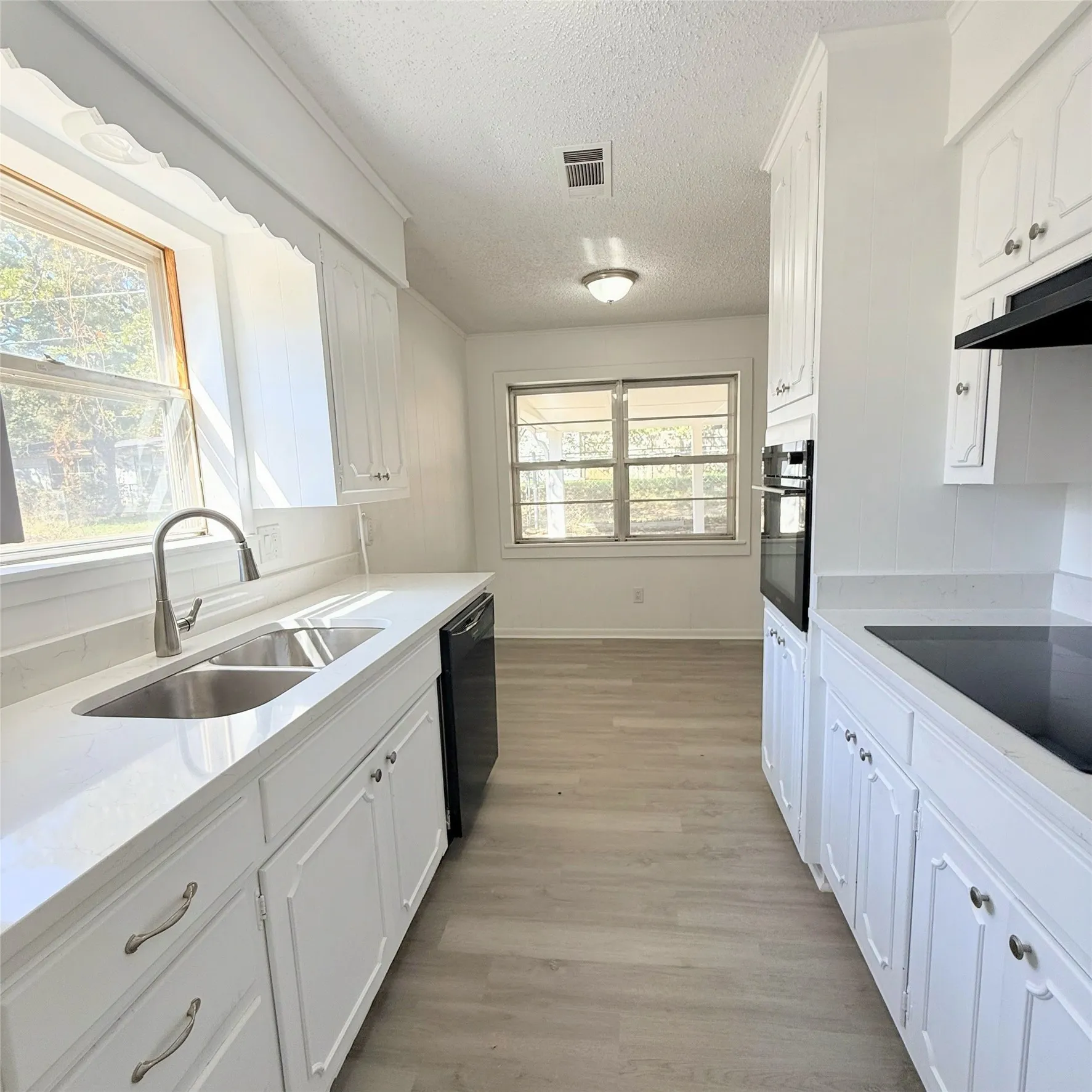 Galley kitchen featuring white cabinetry with hardware accents, stainless steel sink with gooseneck faucet, black built-in appliances including wall oven and cooktop, wood-finish flooring, and abundant natural light from dual windows overlooking the exterior