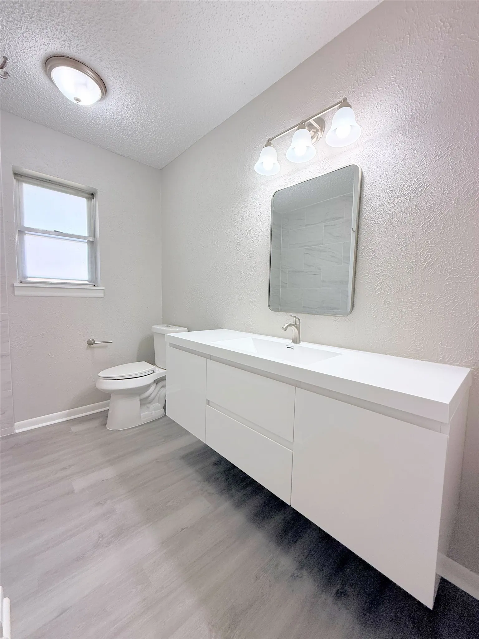 Floating vanity with integrated sink, modern rectangular mirror, overhead vanity lighting, wood-finish flooring, and a window