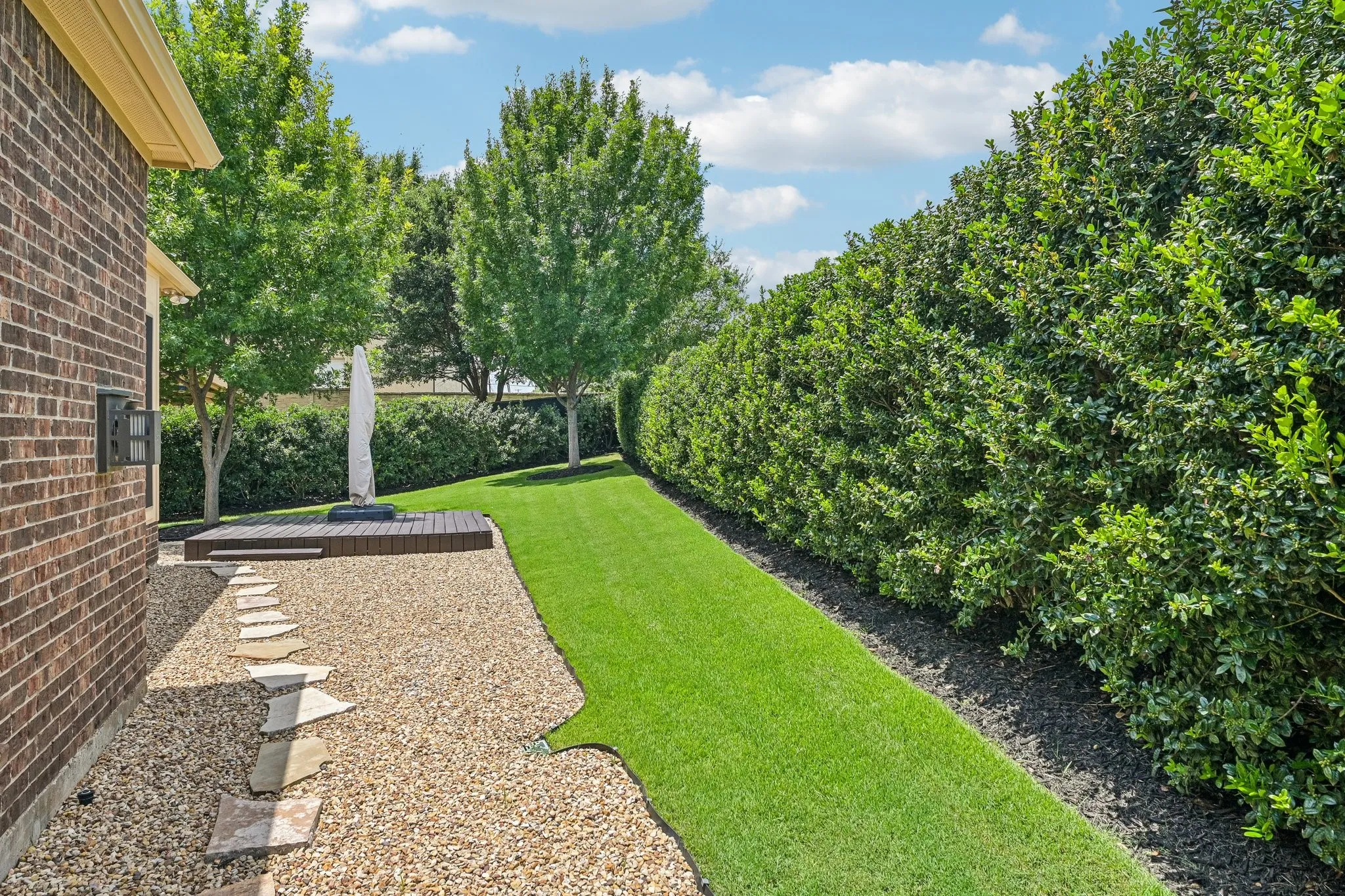 Expansive side yard featuring a manicured lawn, gravel path with stepping stones, a raised wood deck, and mature privacy hedging