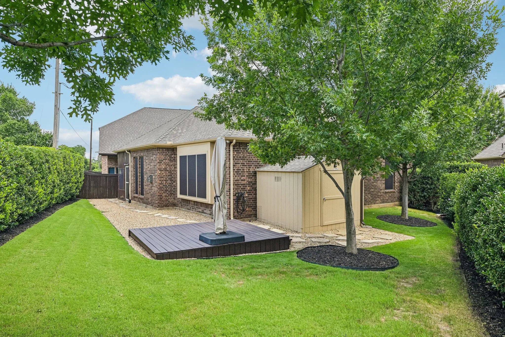 Brick exterior residence showcasing a sprawling rear lawn with mature trees, a composite deck with integrated fire pit feature, and manicured landscaping with defined planting beds and hedging