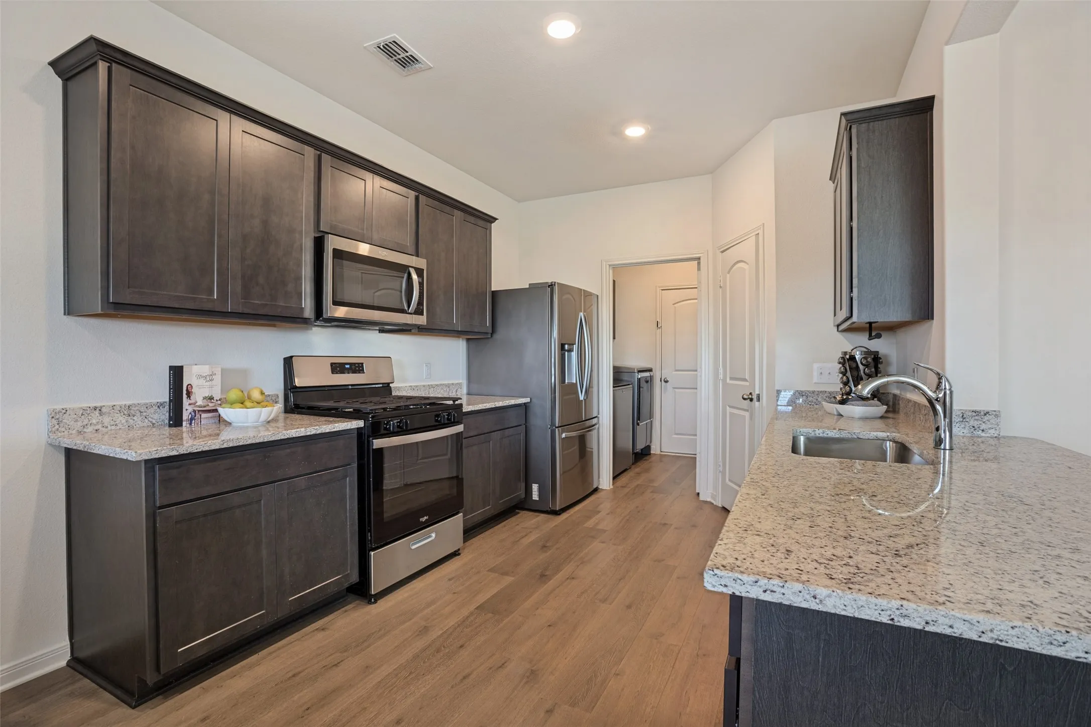 Modern kitchen featuring dark cabinetry with stainless steel appliances, granite countertops, a spacious island with seating, recessed lighting, and wood-finish flooring throughout the open layout