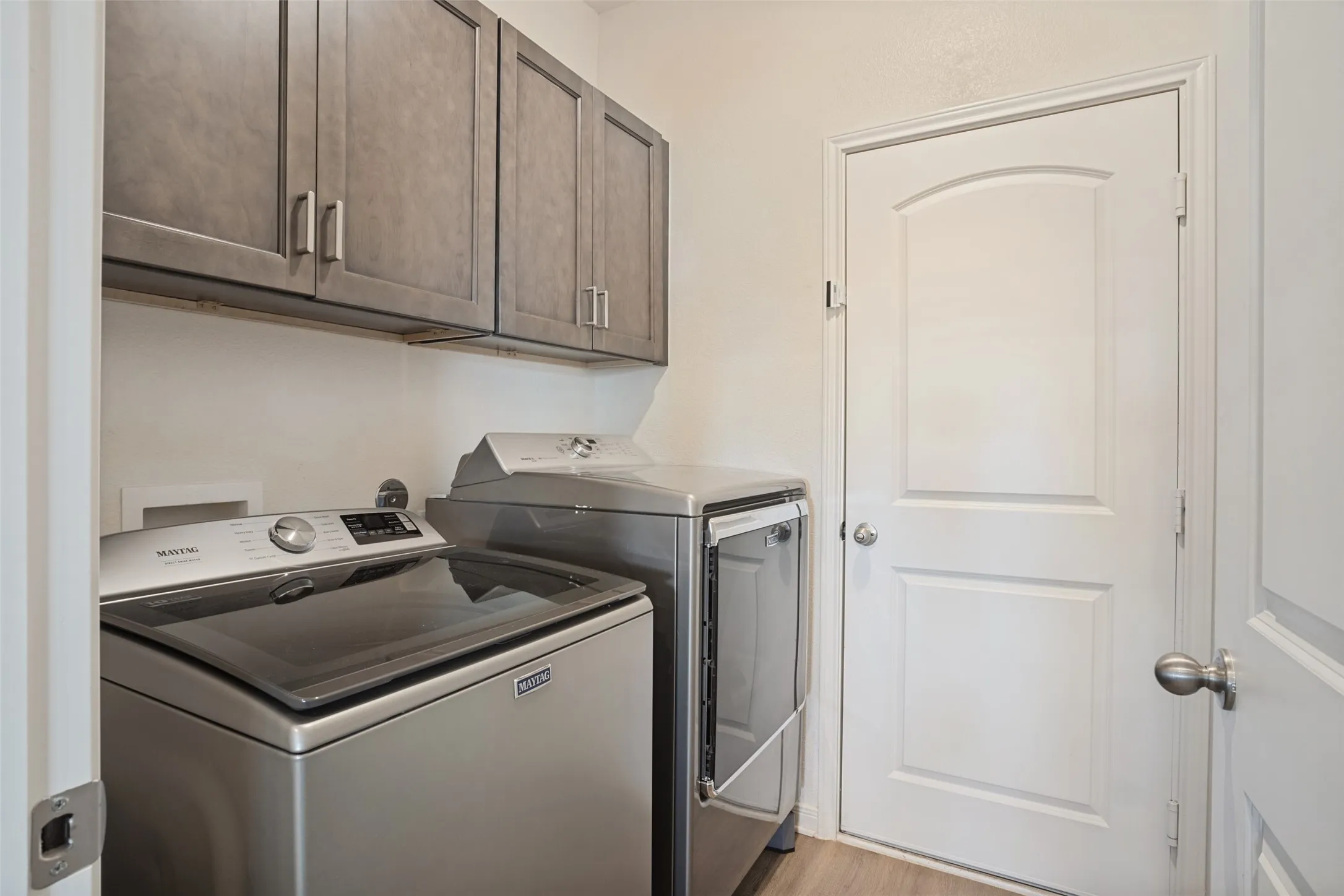 Laundry area featuring wood-finish flooring, overhead cabinetry with brushed nickel hardware, and an arched panel door