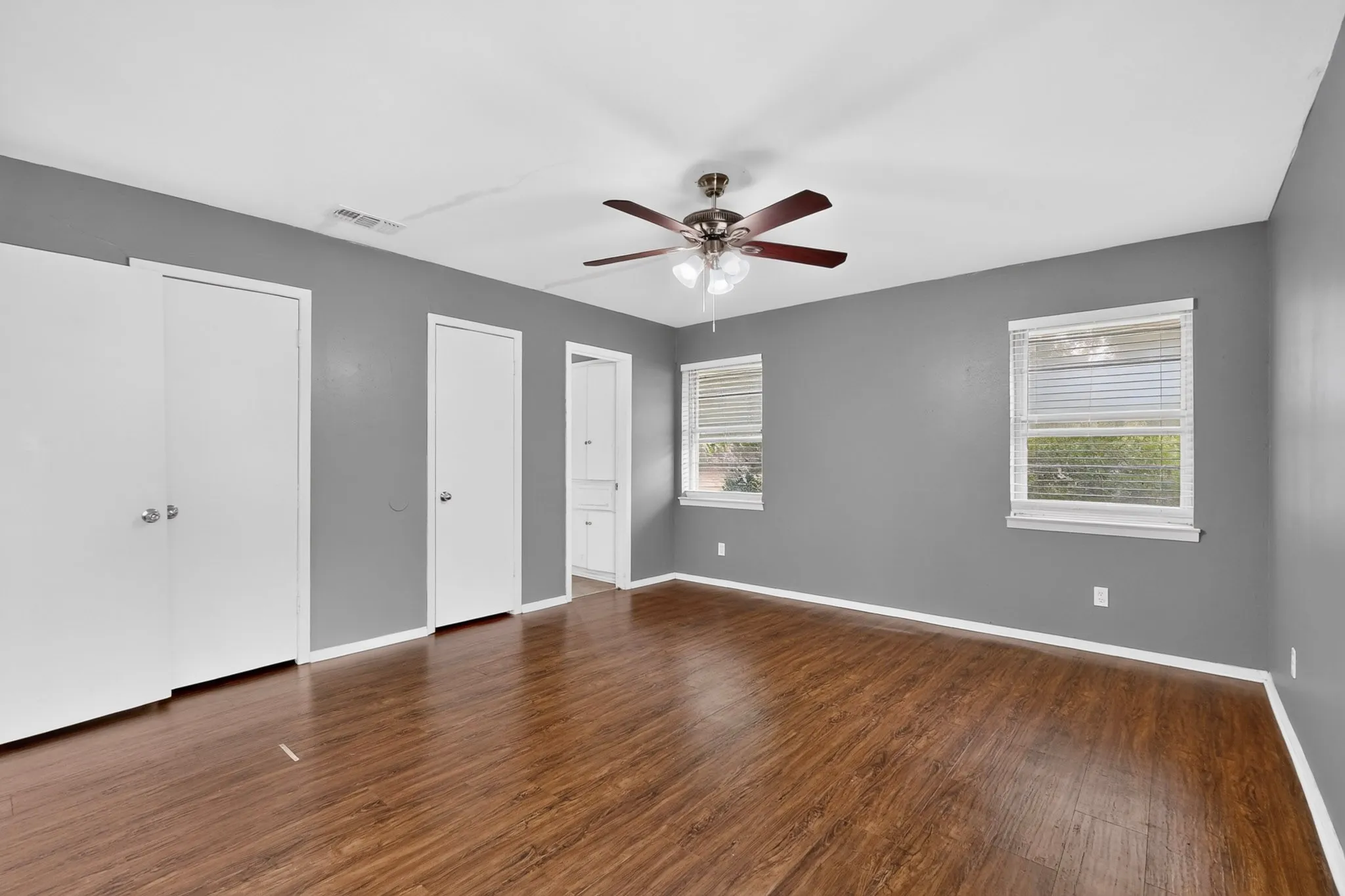 Spacious bedroom showcasing warm wood-finish flooring, ceiling fan with integrated lighting, and multiple white-painted closet doors