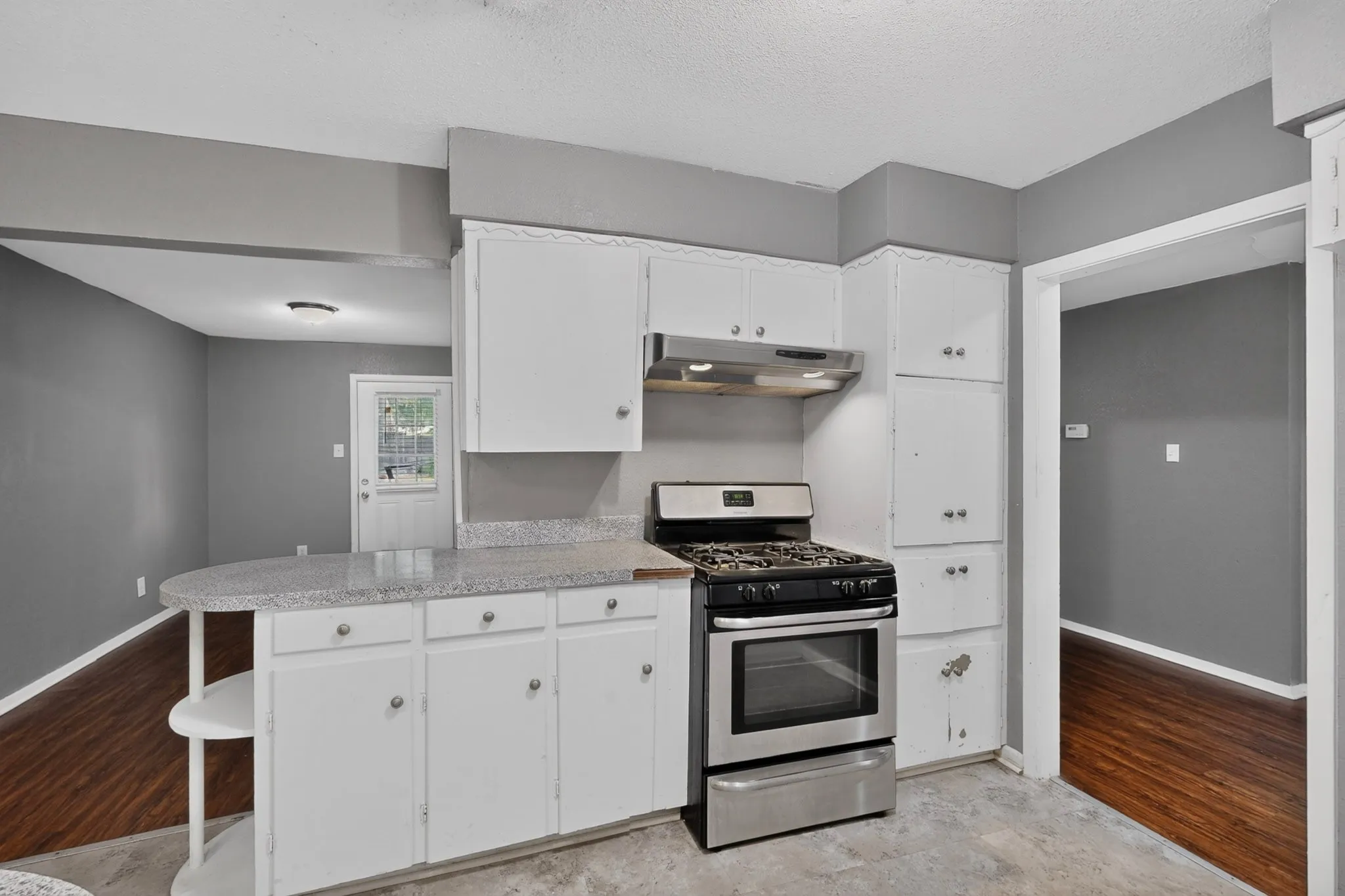 Modern kitchen featuring white cabinetry with ample storage, stainless steel gas range with convection oven, stainless steel range hood, granite countertops, and wood-finish flooring throughout the open layout