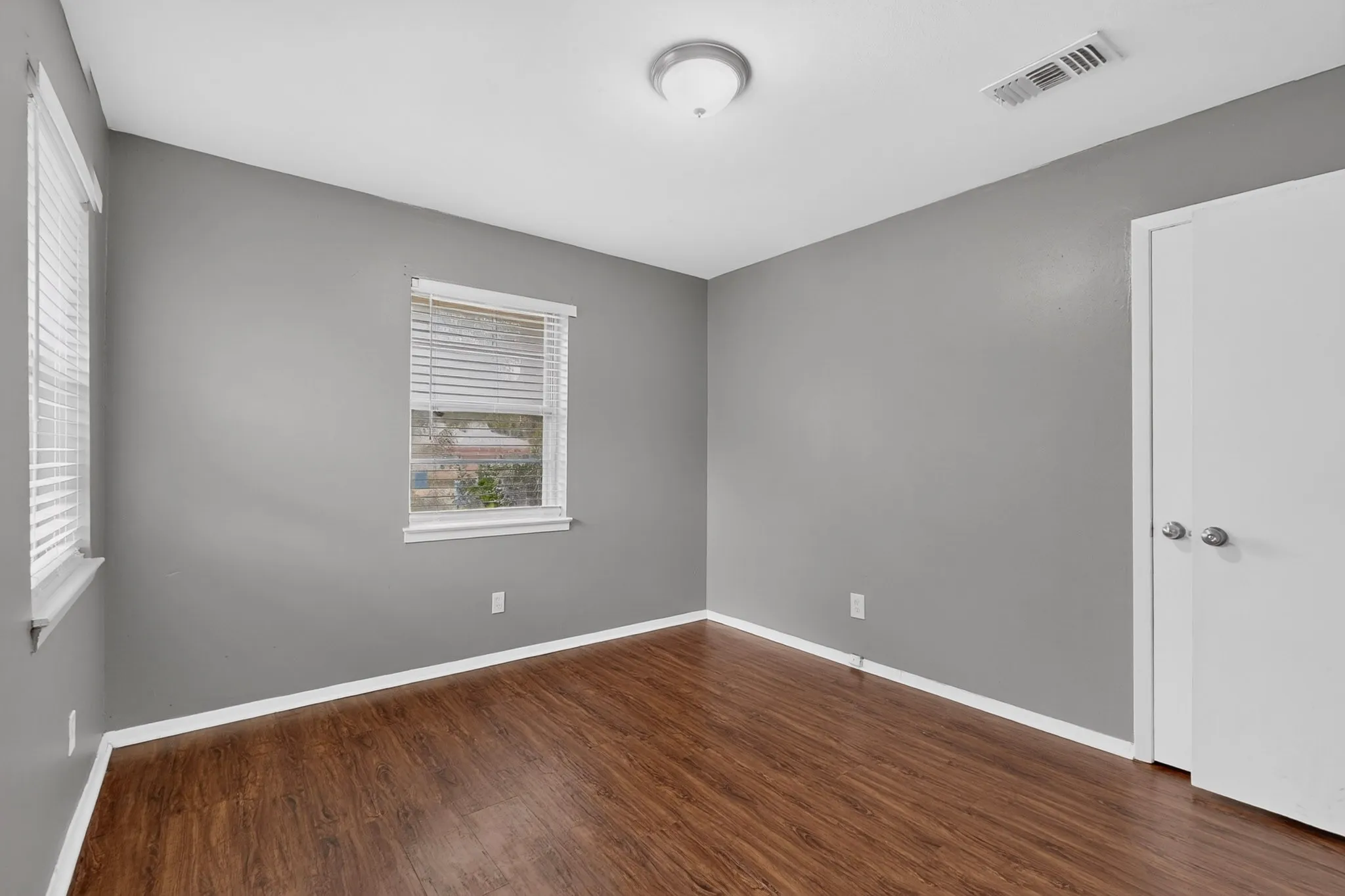 Wood-finish flooring, neutral gray wall tones, white trim, two windows with blinds, and a ceiling-mounted light fixture