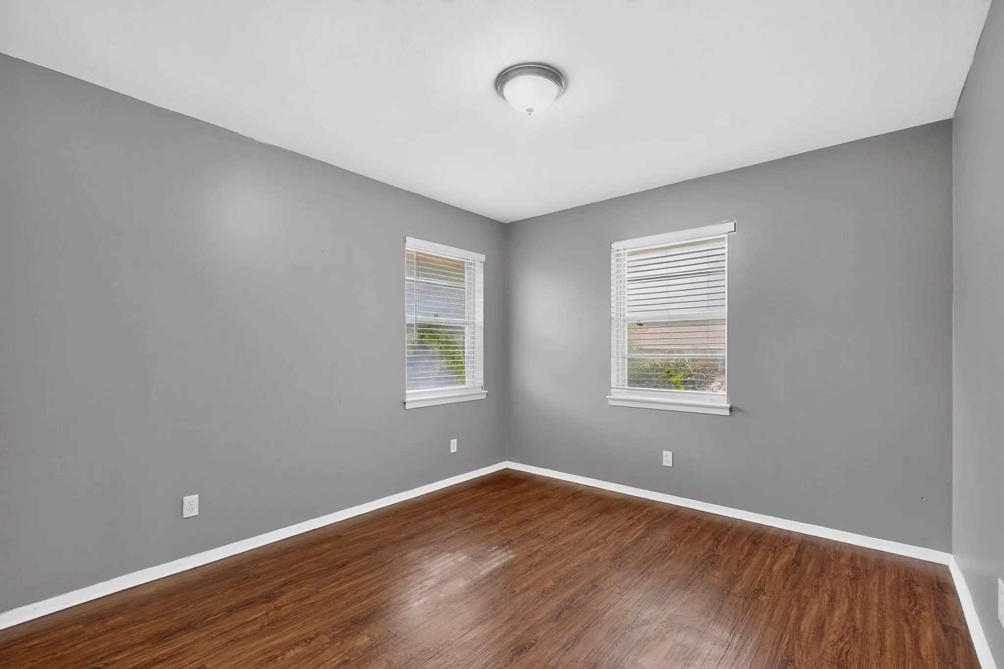 Wood-finish flooring with white trim, two windows with blinds, and neutral grey walls