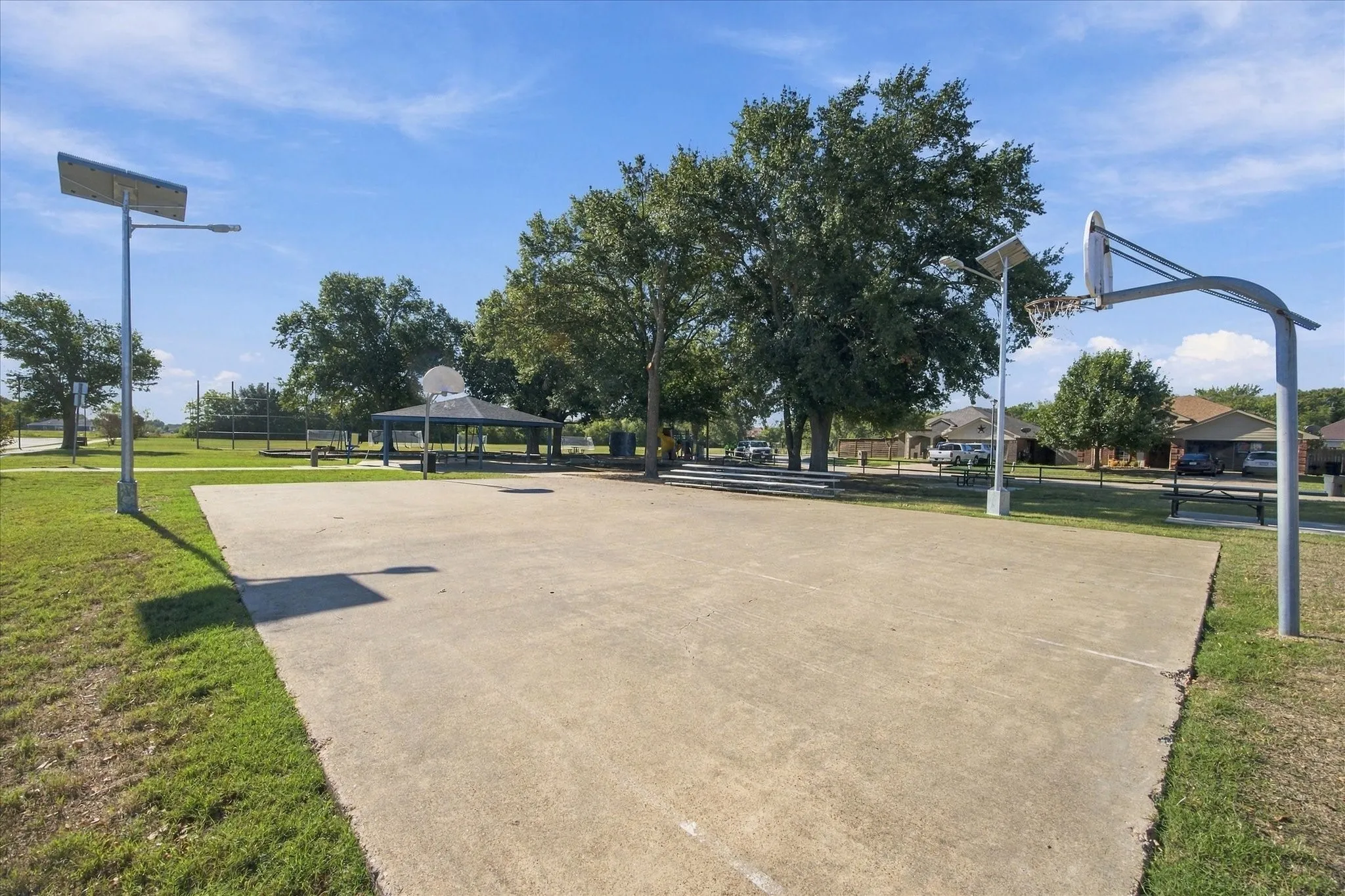 Concrete basketball court, two basketball hoops, multiple mature trees, covered gazebo, and stadium seating
