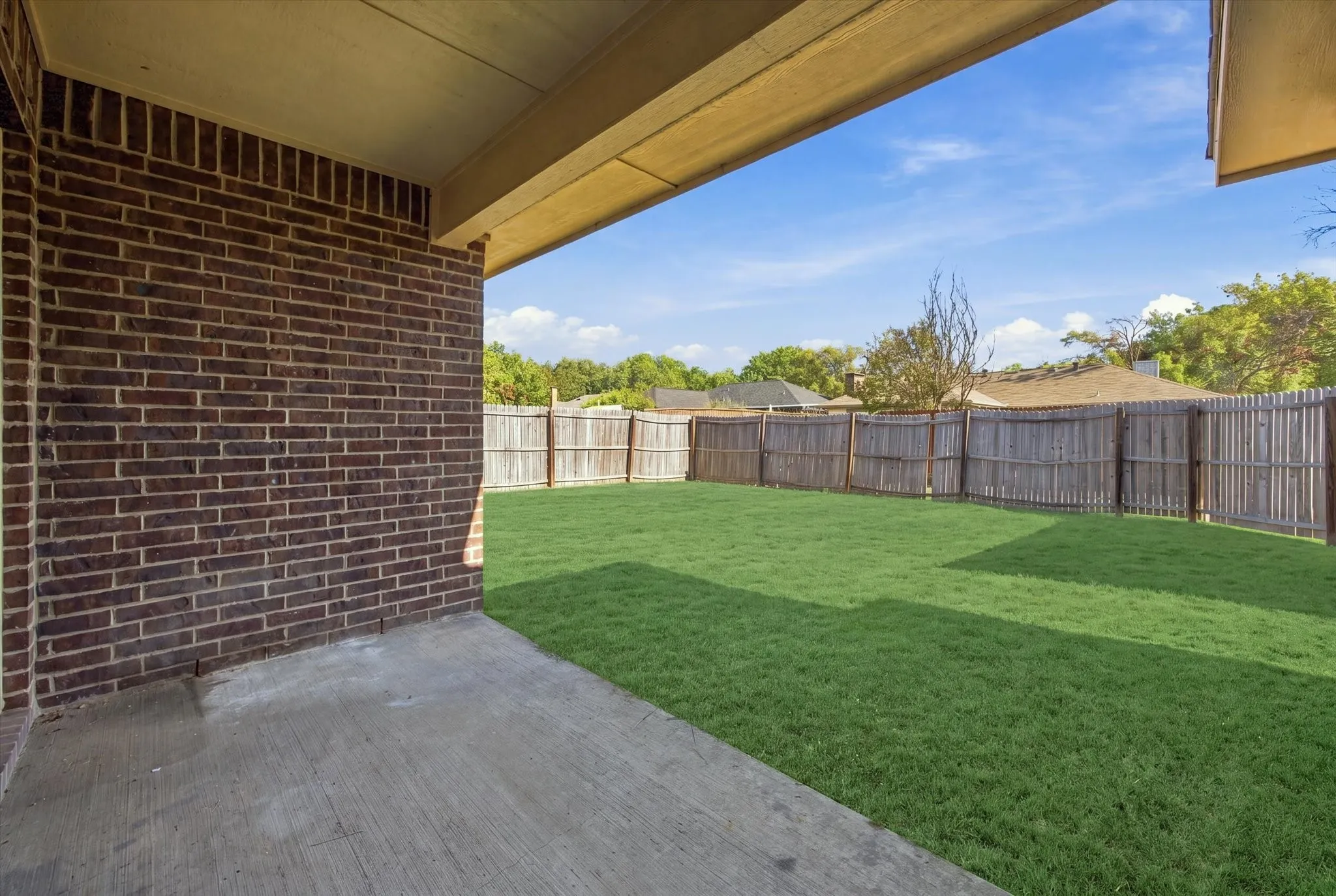 Covered patio with a concrete surface, brick exterior wall, and wood fence enclosing a grass lawn