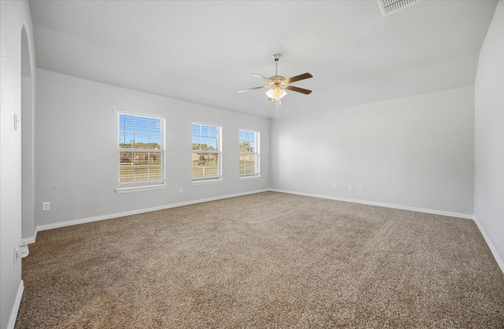 Spacious room featuring neutral carpet flooring, three windows with horizontal blinds, a ceiling fan with integrated lighting, and light-toned wall paint