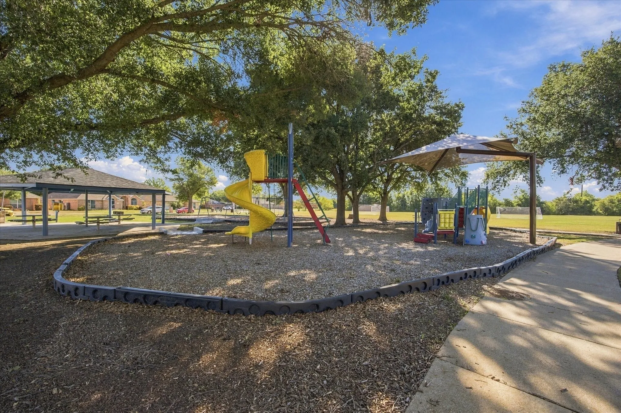 Community park featuring multiple playground structures with slides and climbing equipment, shaded seating areas with umbrellas, mature trees providing natural canopy coverage, and rubber safety surfacing throughout the recreational grounds